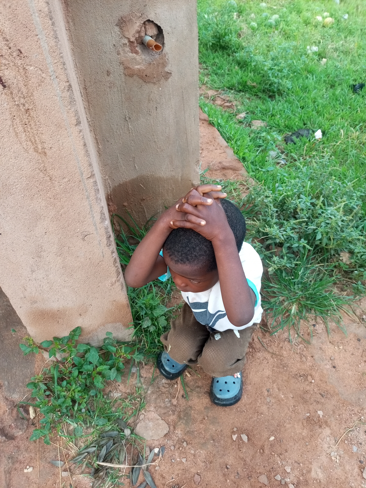 A young child is crouched on reddish-brown dirt next to a concrete structure. The child, wearing a white t-shirt with a black and gray striped pattern and brown checkered pants, has their hands clasped over their head, appearing to be in distress or covering their ears. Blue clogs are on their feet. Behind the child, green grass and some scattered leaves are visible. A small, partially visible pipe protrudes from a hole in the concrete structure. The overall setting suggests an outdoor, possibly rural, environment.