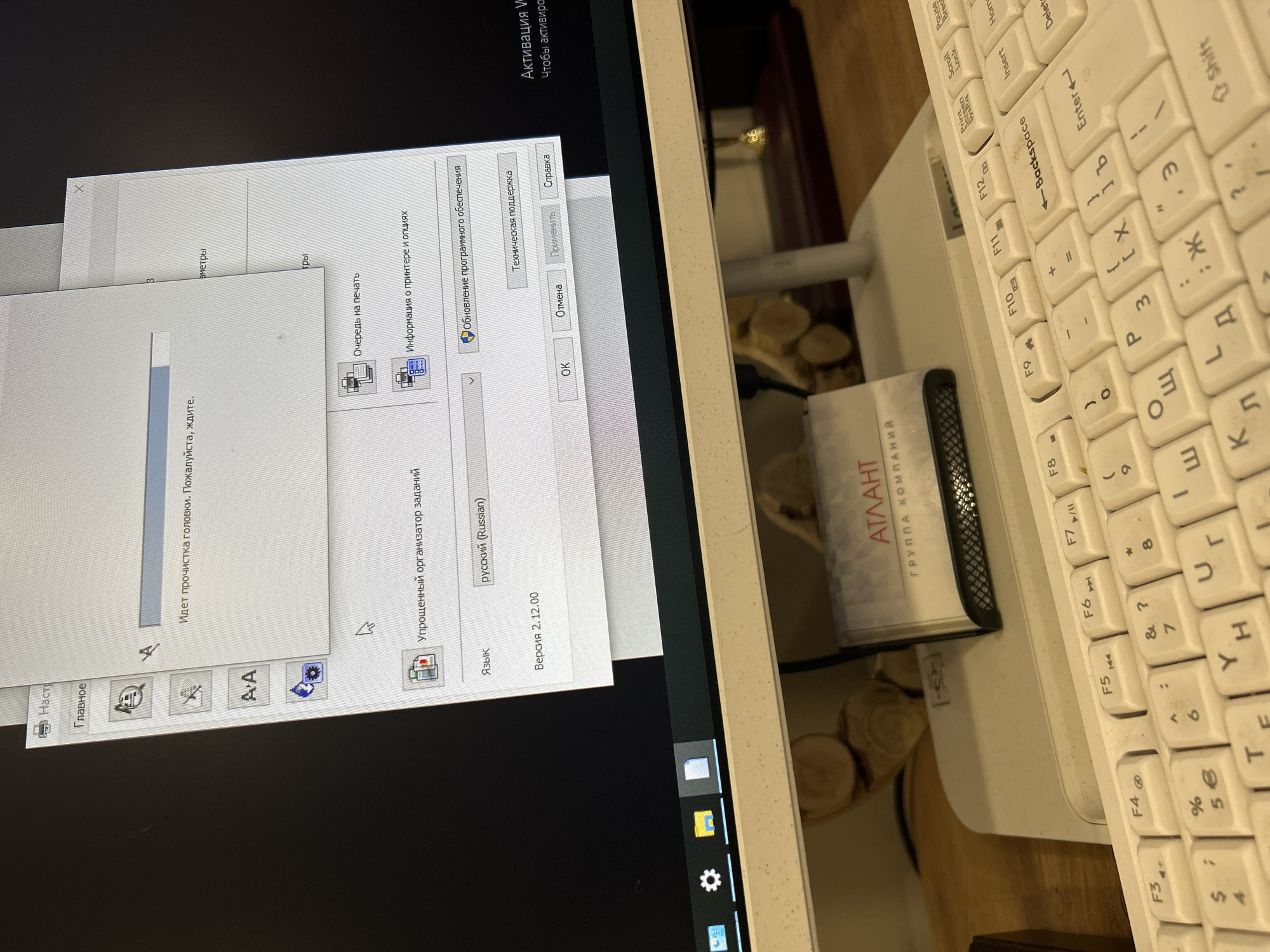 The image shows a computer screen displaying software in Russian, including a window that says "Идет прочистка головки. Пожалуйста, ждите" which translates to "Head cleaning in progress. Please wait." The software appears to be related to printer management. The language selected is "русский (Russian)." Next to the screen is a white keyboard, with Russian characters on the keys. A business card holder is located in front of the keyboard with the word "АТЛАНТ" ("ATLANTA") in Russian and "ГРУППА КОМПАНИЙ" ("GROUP OF COMPANIES") written on it. The location in the city of Kemul, Russia is not possible to determine from the image.