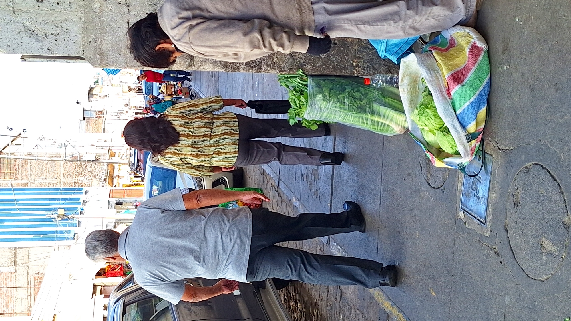 Three people are standing on a city street next to a car. A bag of lettuce and a bag of celery are on the ground.