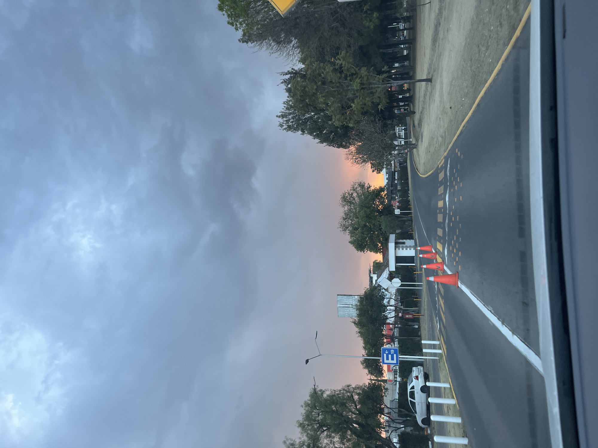 This image depicts an outdoor road scene observed from the interior of a vehicle, facing forward, at approximately dusk or dawn. The location is identified as being near the Universidad de las Américas Puebla (UDLAP) in San Andrés Cholula, Mexico, evidenced by the "E 19" parking sign which corresponds to campus designations.
The sky is predominantly covered with dark gray, dense clouds, with a striking orange-pink hue visible along the horizon line, indicative of either sunrise or sunset conditions.
The foreground is dominated by an asphalt road, gently curving towards the right. A solid white line demarcates the left edge of the visible lane, while a solid yellow line marks its right edge. Across the road, a series of yellow rectangular speed bumps or rumble strips are present, accompanied by smaller yellow reflective road markers. Four orange traffic cones are positioned in a linear arrangement on the road, partially within the left lane.
To the left of the road, a row of white cylindrical bollards is visible. Beyond these, a white four-door sedan is parked. A gray street light pole with two luminaires stands prominently. Beneath the street light, a blue square sign displays a white "E" followed by "19" and a left-pointing arrow, designating a parking zone. Further left, partially obscured by trees, a tall building with a metallic facade rises in the background. A small, white, single-story structure resembling a guardhouse with a brown roof is also present on the left, near two white circular signs mounted on posts. The left roadside is heavily vegetated with various trees and shrubs.
To the right of the road, a wide strip of dry, light-colored grass extends, bordered by a dense line of trees, including both evergreen and deciduous species. A yellow triangular warning sign is partially visible on the extreme right edge of the frame. No individuals are present, and no active movements or interactions are depicted in the scene.