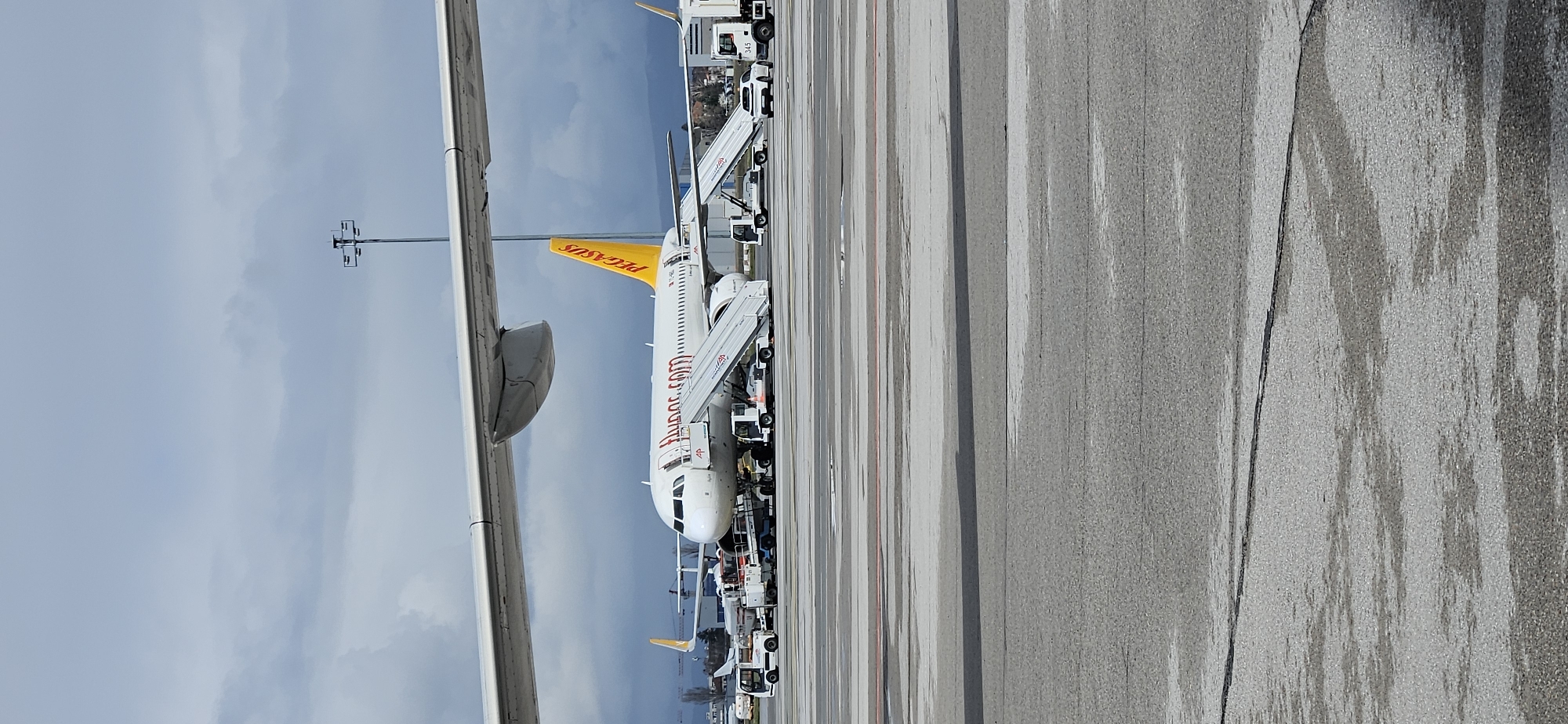 The image depicts a Pegasus Airlines aircraft on the tarmac at what appears to be an airport. The aircraft is white with a distinctive yellow tail featuring the "PEGASUS" logo. The fuselage has "flypgs.com" painted in red. Passenger stairs are attached to the aircraft, and service vehicles are lined up next to it. The tarmac is wet and shows signs of recent rain. Another aircraft can be partially seen in the background.
The presence of Pegasus Airlines and the general airport setting would potentially indicate that the location is Sofia Airport (SOF) in Sofia, Bulgaria, as Pegasus Airlines operates flights to and from Sofia.