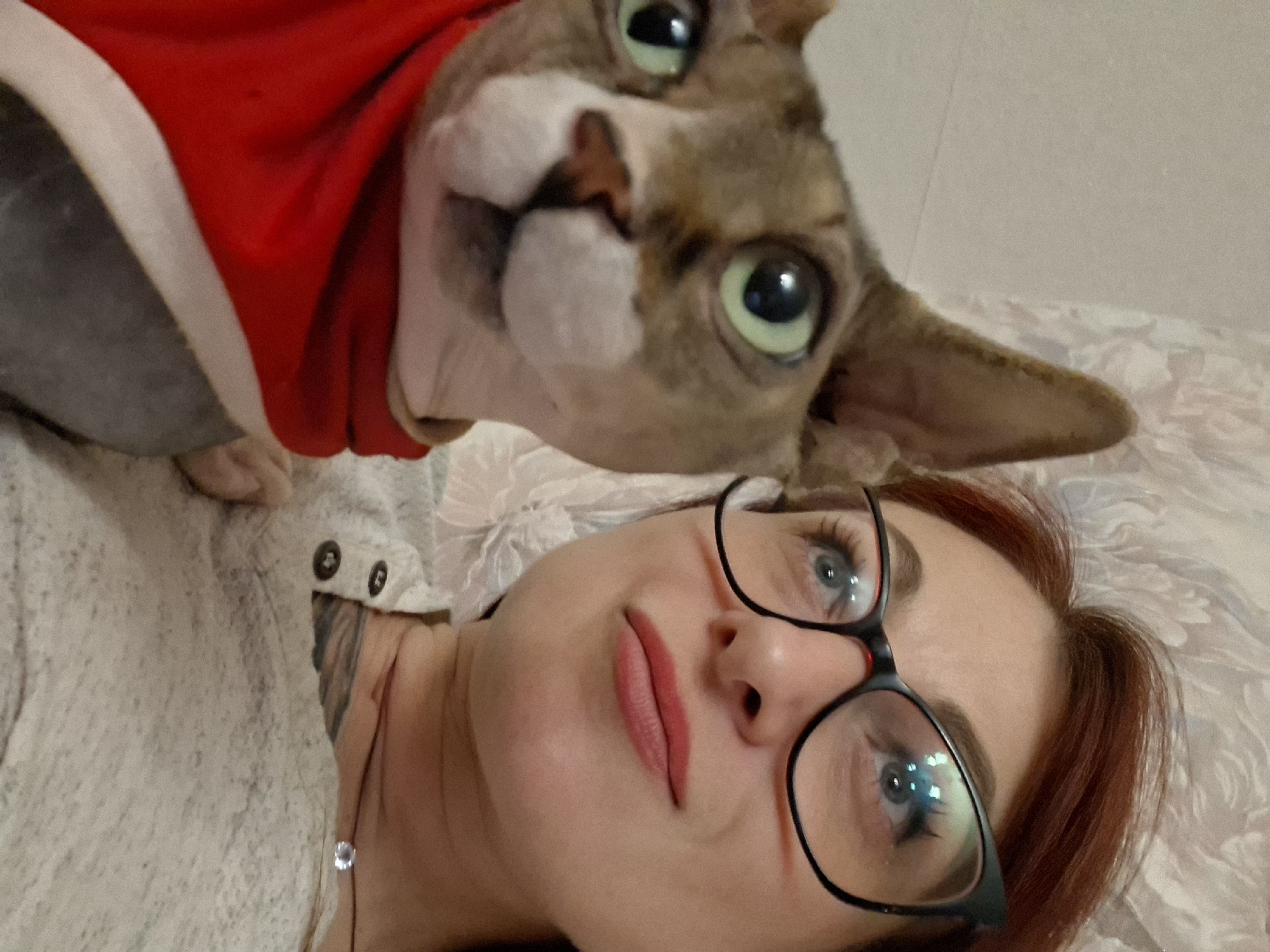 This is a close-up, indoor, selfie-style photograph taken from a slightly high angle. In the foreground on the left is a Sphynx cat, wearing a red garment that covers its body. The cat has light brown and grey tabby markings on its face and large, green eyes. To the right of the cat is a woman wearing black-rimmed glasses with a slight upward curve at the edges. She has auburn hair and is looking up and to her right with a slight smile. She is wearing a light grey, button-up shirt. The background appears to be a bed with a patterned comforter. The scene is likely in a domestic setting within Забалоцкі сельскі Савет, Belarus.