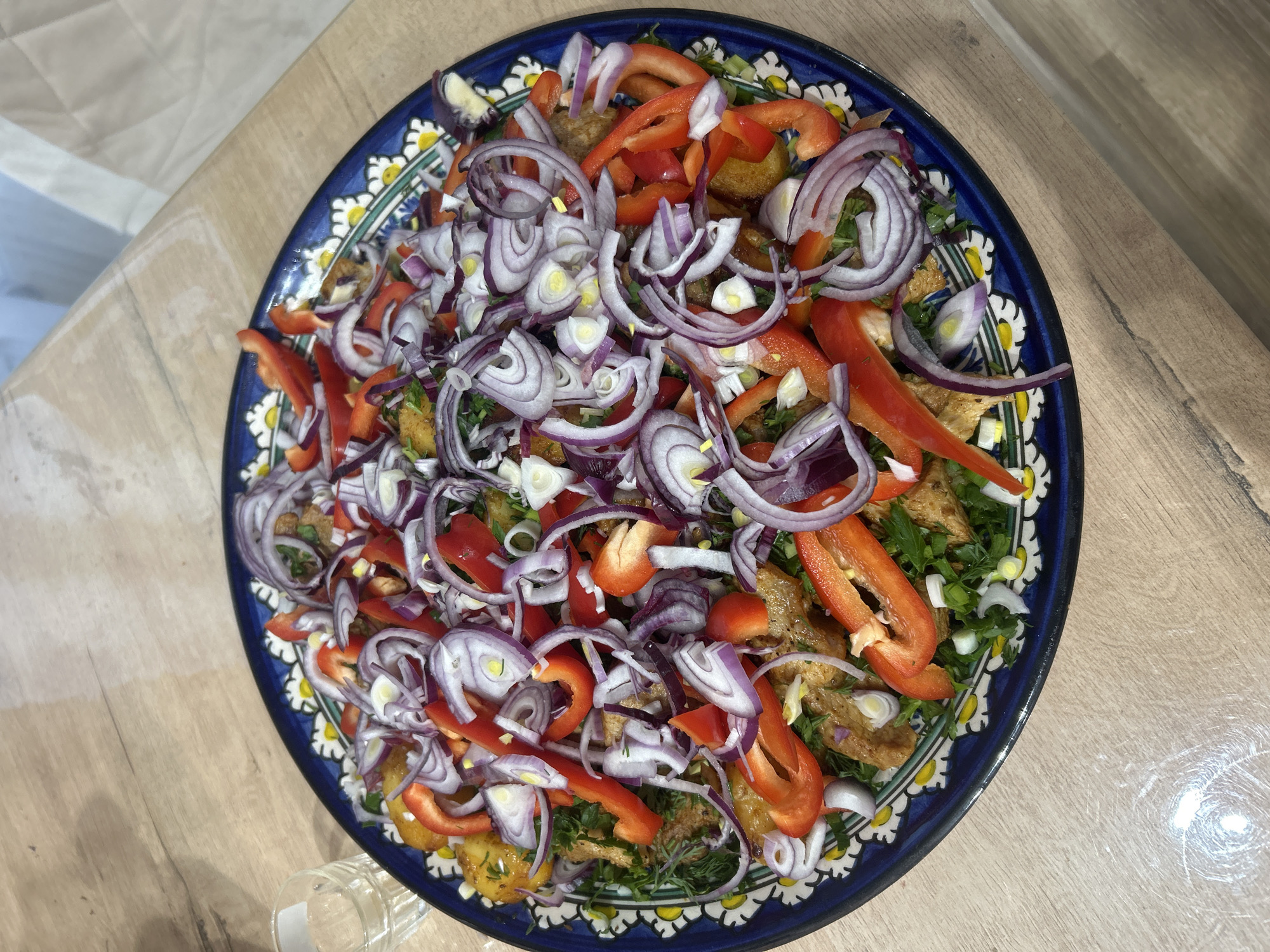 A high-angle shot shows a platter of food, possibly a salad, resting on a light-colored wooden table. The platter is dark blue with a white floral pattern around the rim. The salad consists of red onion rings, sliced red bell peppers, chopped greens (possibly herbs), and what appears to be fried potatoes or meat. A portion of a clear glass is visible in the lower left corner. The specific location in Chaikovsky, Russia, cannot be determined from this image.