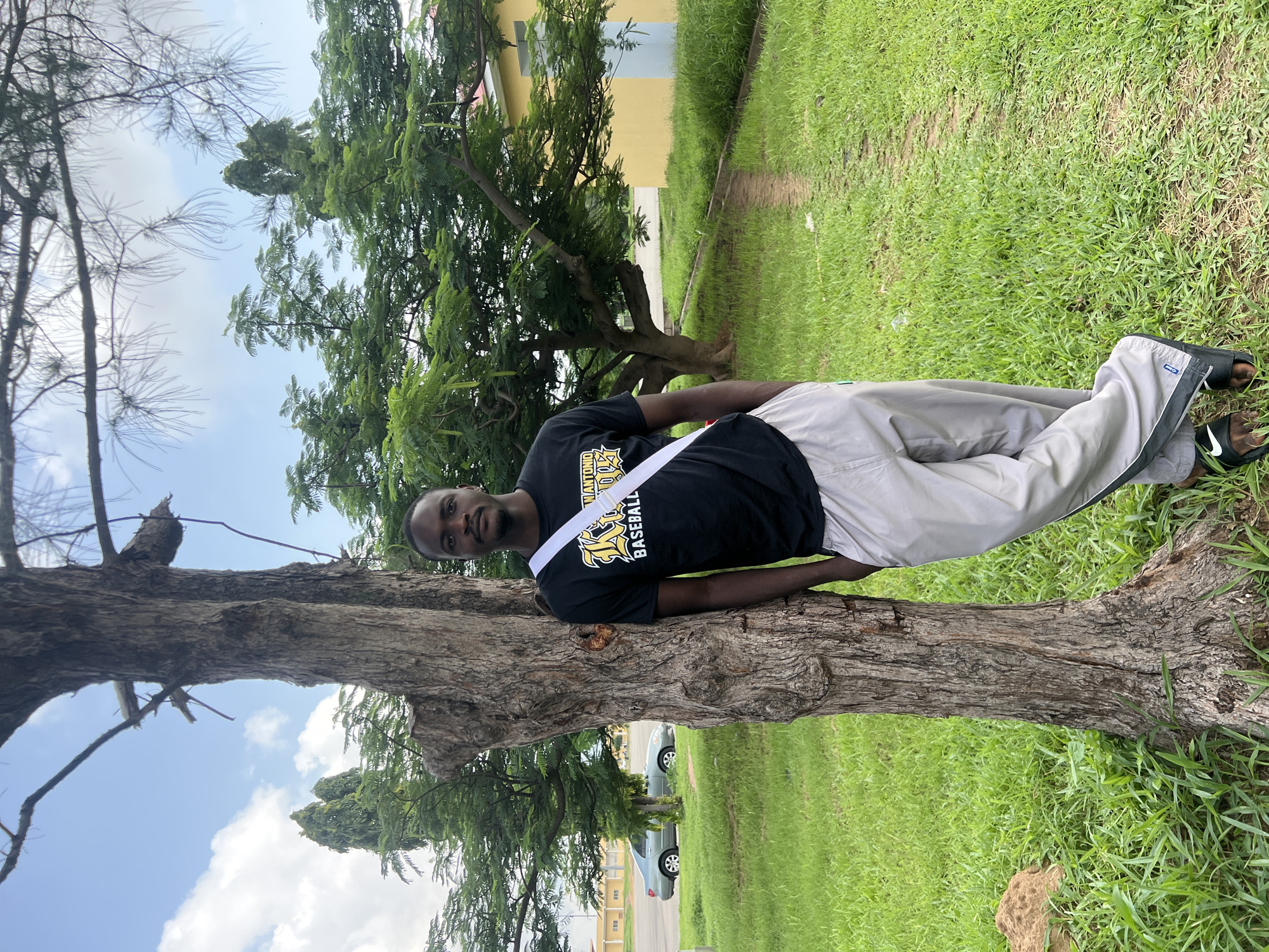 A young Black man is lying down, resting against the trunk of a large tree. He is wearing a black t-shirt with a yellow and white logo, light gray athletic pants with a dark stripe down the side, and black slide sandals. A white strap runs diagonally across his chest. The man is looking towards the camera with a neutral expression.
The setting appears to be a park or outdoor area with lush green grass and trees. In the background, a light yellow building with windows can be seen, along with a paved pathway. A silver car is parked on the side of the pathway. The sky is visible with some clouds, suggesting it is daytime. The overall impression is a casual and relaxed moment.
The visible text on the man's shirt reads "ANTONIO BASEBALL".