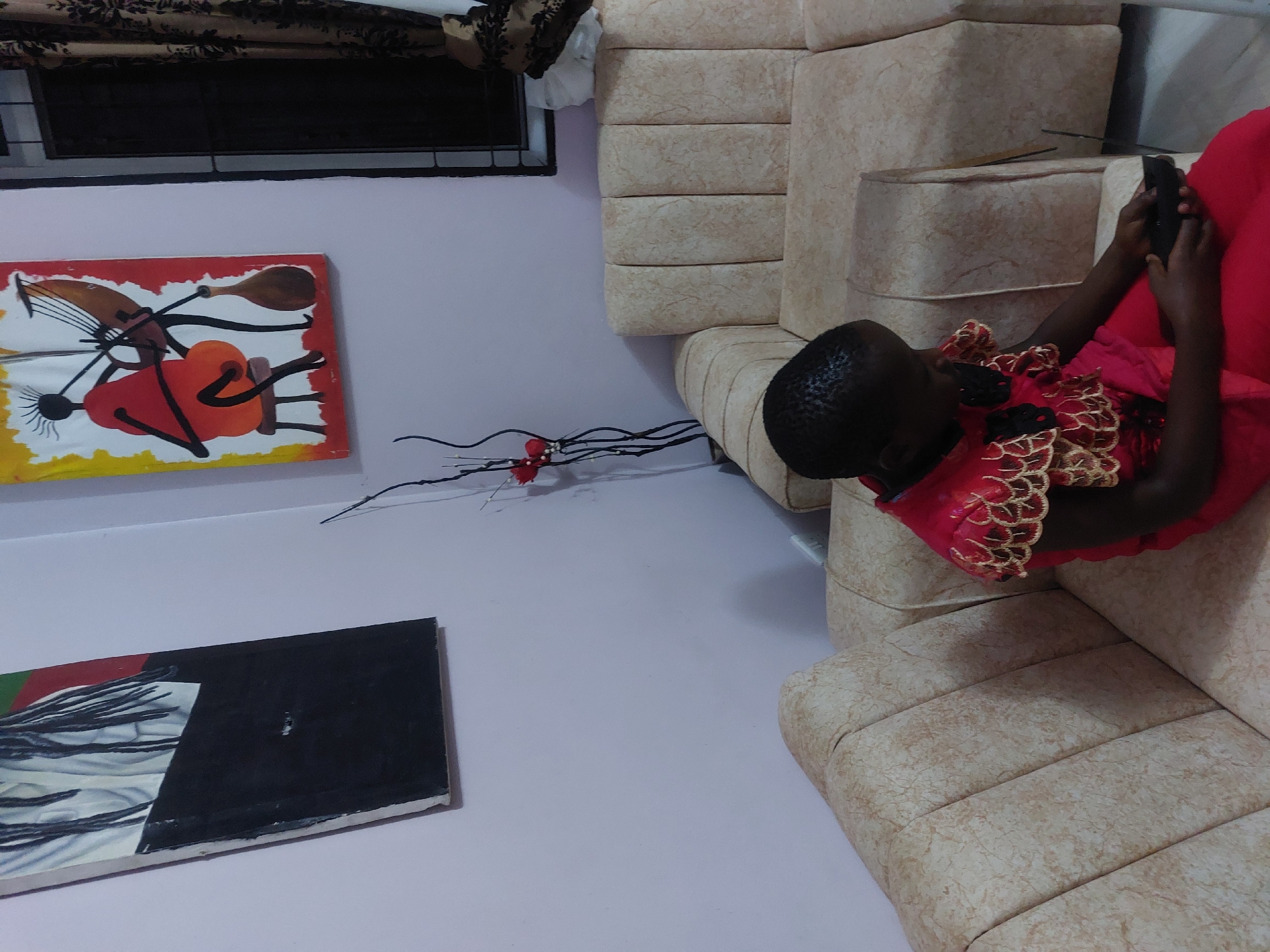 A child is lying on a tan, quilted sofa, looking at a black phone in their hands. The child is wearing a red dress with gold trim. A black window frame with curtains is in the upper left of the shot. Two paintings are on the wall: an abstract painting with black, red, yellow, and white, and a black and white painting. A decorative spray with red flowers sits against the wall. The location is possibly a home in Dar es-Salaam, Tanzania.