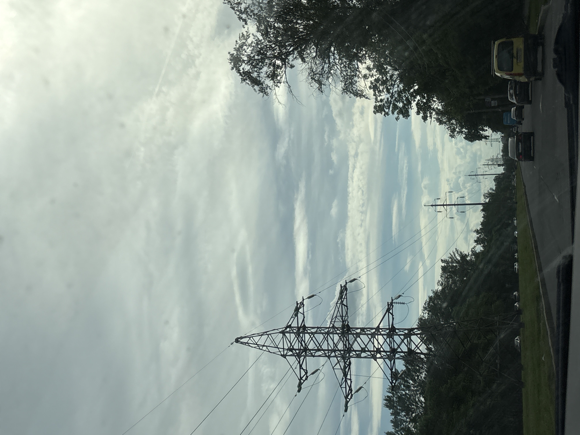 The image presents a daytime view from the interior of a vehicle looking forward through the windshield, depicting an urban or suburban road in Saint Petersburg, Russia, under an overcast sky. The sky is primarily covered by a textured layer of stratocumulus clouds, exhibiting brighter regions towards the right and a subtle, linear contrail visible in the upper right quadrant.
A large, dark lattice-steel electricity transmission pylon stands prominently on the left side, extending from the lower-left into the mid-upper portion of the frame. Multiple high-voltage power lines emanate from this pylon, traversing horizontally across the sky. Below the pylon and continuing towards the left is a dense expanse of green trees and foliage. A grassy median strip is visible at the base of these trees, where several white passenger cars are partially obscured.
On the right side of the image, a dense, dark treeline of deciduous trees forms a silhouetted boundary along the road, extending into the distance. Further down this side, additional, smaller utility poles carrying power lines are discernable.
The center and lower sections of the image are dominated by a multi-lane asphalt road, receding into the background. Several vehicles are observed in traffic, moving away from the viewer. These include a dark sedan in the immediate foreground, a blue commercial van (consistent with a GAZelle) further ahead, and a yellow commercial van (resembling a Citroën Berlingo) positioned on the far right side of the road or shoulder. The lower edge of the image frames a portion of the vehicle's dashboard or windshield base. No individuals are visibly identifiable.