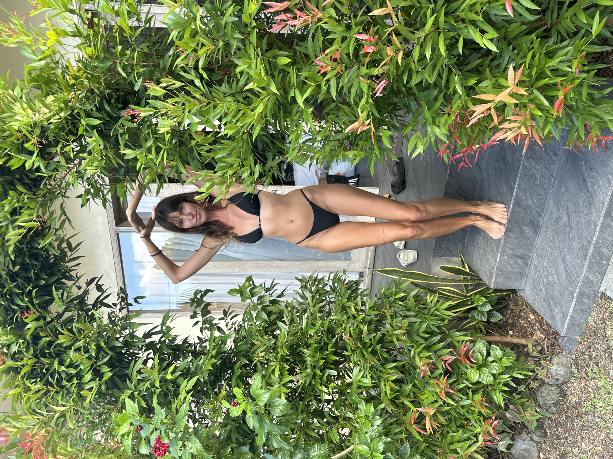 A woman in a black bikini stands on gray tiled steps, partially framed by dense green foliage with red-tipped leaves in an outdoor setting. Her arms are raised, with one hand touching the foliage and the other near a window behind her. The surrounding vegetation includes diverse plant types, and the tiled steps lead to a building with visible windows and white walls. There are plants to the side and base of the steps. The location is North Kuta, Indonesia.
