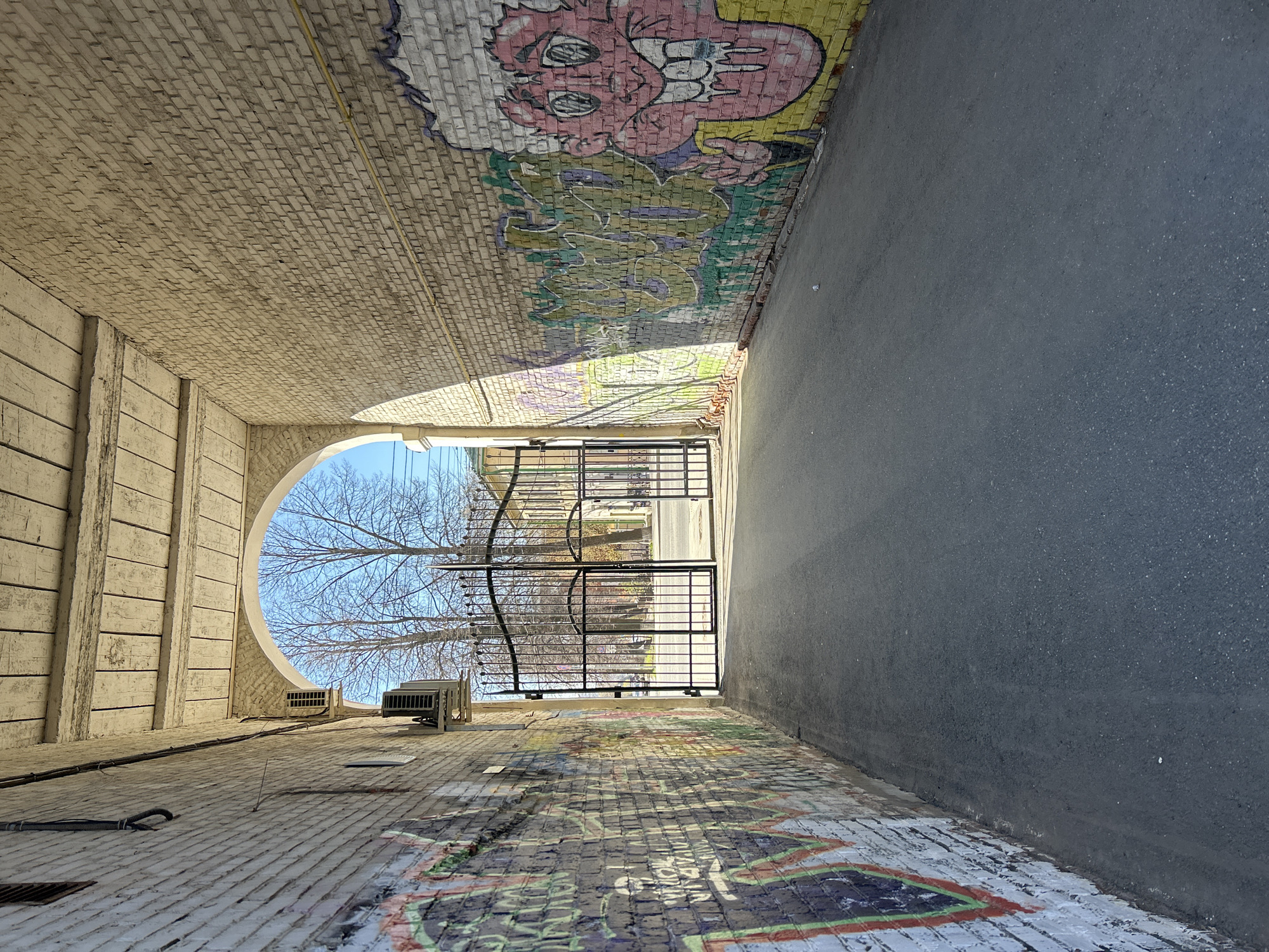 The image shows a narrow passage in Izhevsk, Russia, with brick walls and an arched opening at the end. Graffiti covers the brick surfaces, including a cartoonish figure at the top. A black metal gate partially obscures the view beyond the archway, where trees and a street are visible. The passage floor is a mix of brick and asphalt. An air conditioning unit is affixed to the brick wall near the archway. The image is taken from a low angle, looking towards the end of the passage.