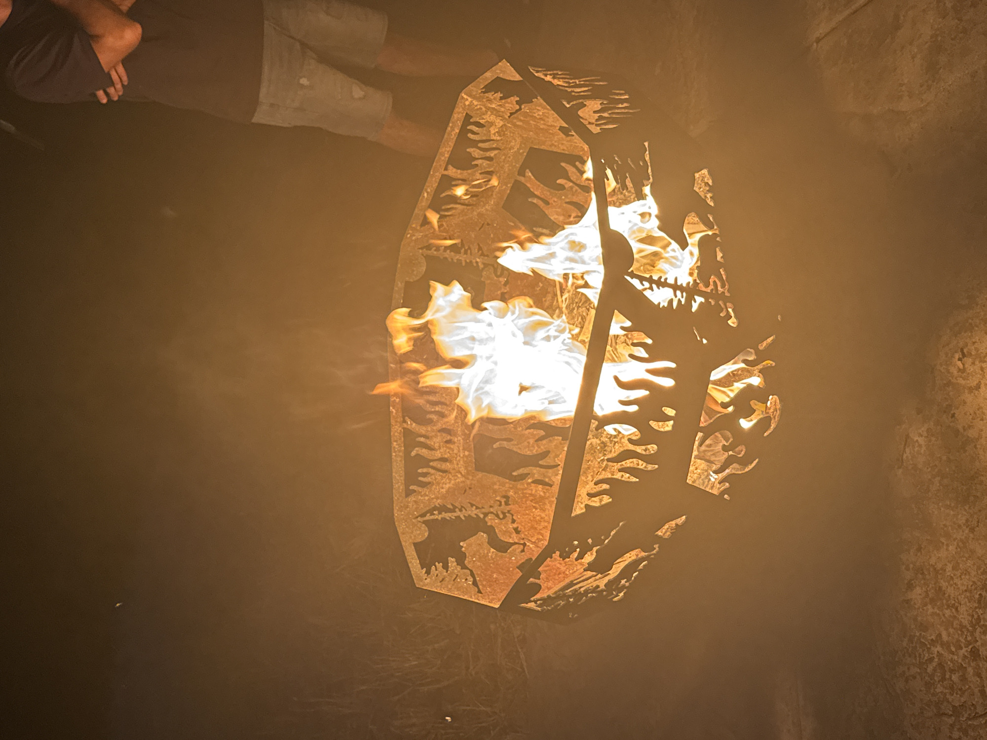 At night, a fire burns in a circular metal fire pit with cut-out designs of trees and animals. The fire pit is on a paved surface. A person's legs and torso are visible on the right side of the frame, wearing a dark t-shirt and denim shorts. The fire is bright white and yellow, with flames reaching upwards. The background is dark and out of focus, with some visible grass and brush.