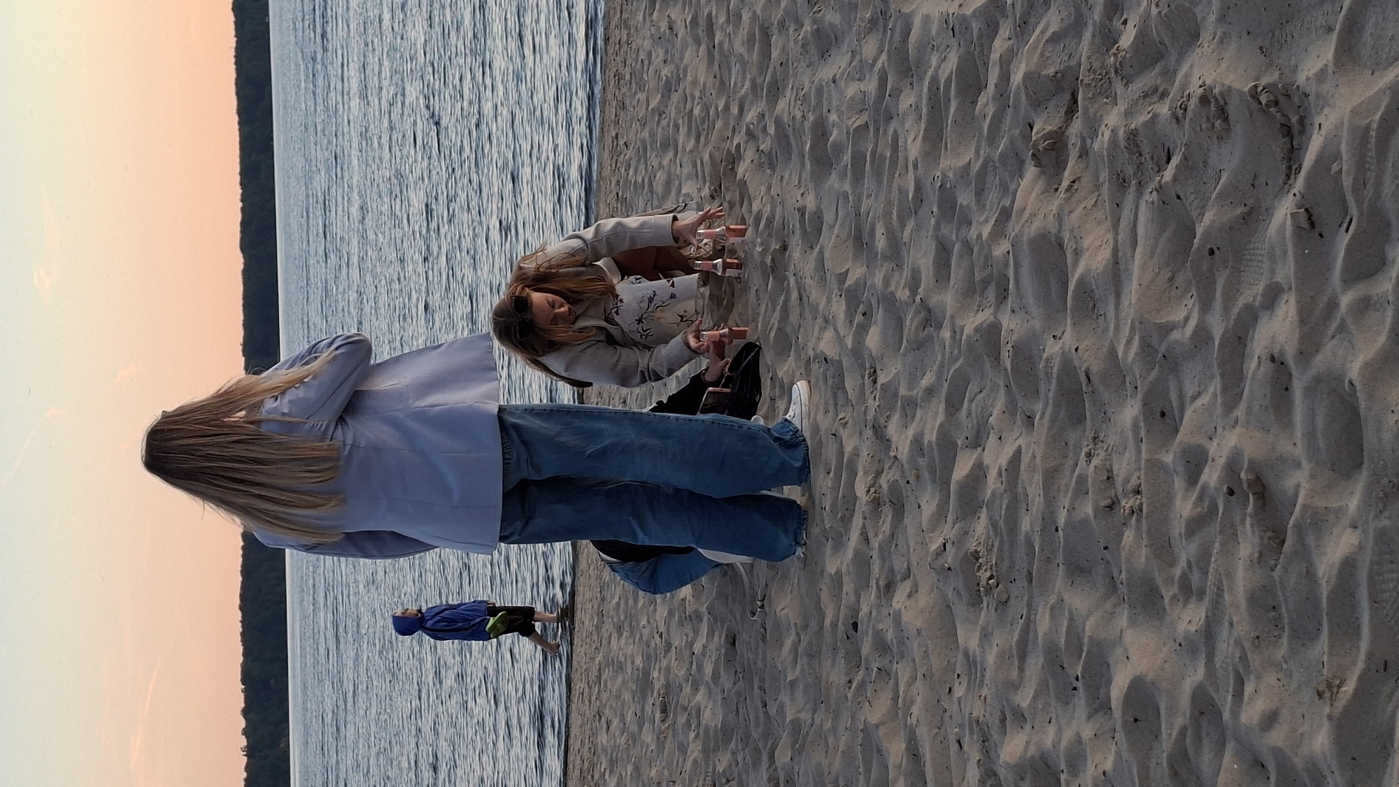 In Tallinn, Estonia, on a sandy beach adjacent to the Baltic Sea, a group of individuals is engaged in an activity near the water's edge. The sky displays a soft gradient of sunset hues, ranging from pale orange to light pink, with a thin band of trees forming a distant treeline.
In the foreground, a person wearing a light blue blazer and blue jeans stands with their back to the camera, facing the water. To their right, a young woman wearing a grey coat over a floral dress kneels on the sand. She is arranging several small, pink bottles in a semi-circle on the beach. Another individual, partially visible and wearing blue jeans, is also kneeling in the sand near the woman with the bottles.
In the background, a child in a blue hooded sweatshirt and black shorts walks in the shallow water, facing away from the viewer. The water is calm and reflects the colors of the sky. The sand is textured with ripples and small indentations. The overall lighting suggests it is late evening.