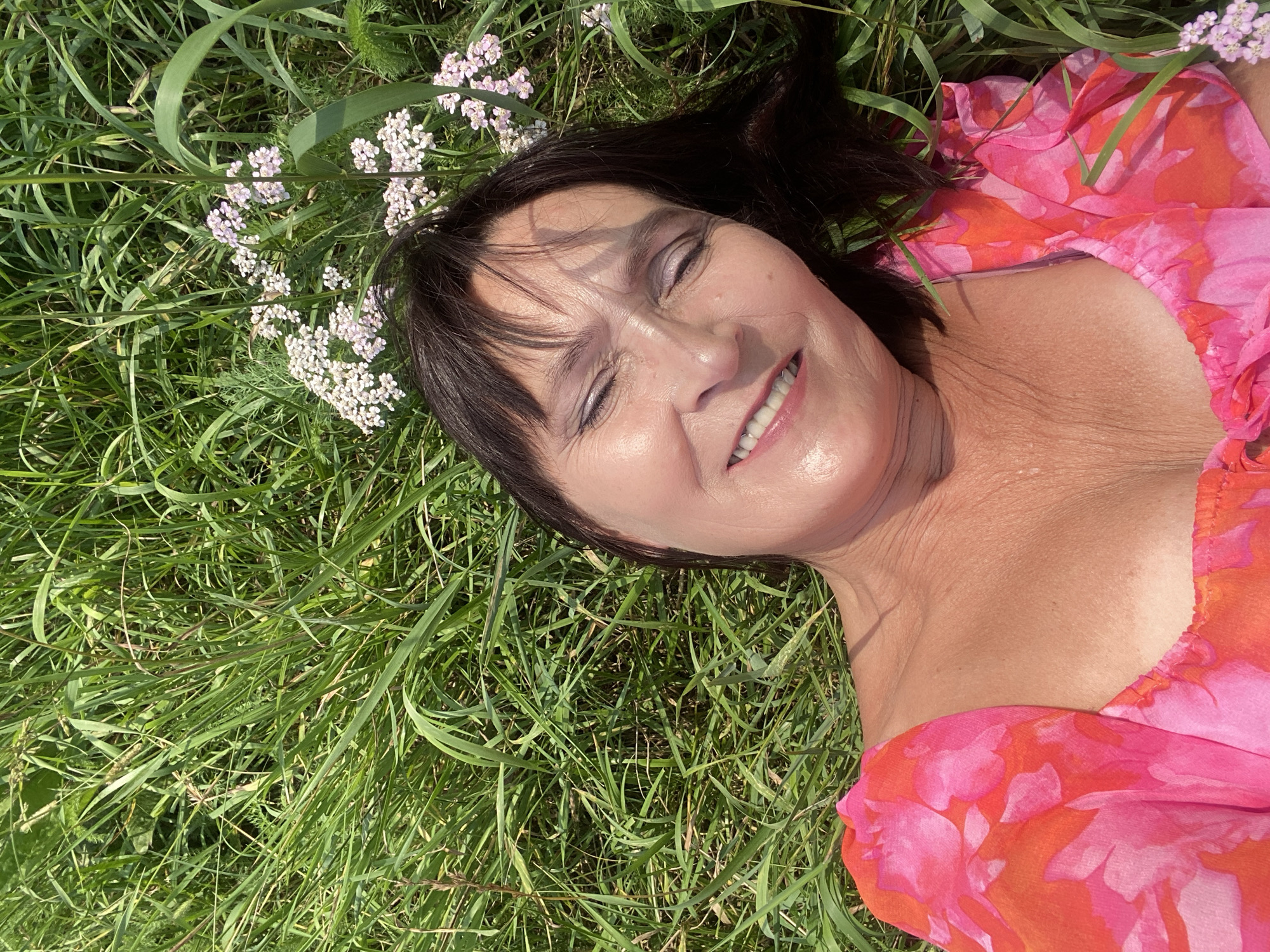 A middle-aged woman with dark hair lies on her back in a grassy field, her eyes closed and a smile on her face. She is wearing a pink and orange floral print top with a low neckline. The grass is tall and green, with some small white and pink wildflowers scattered throughout. The image appears to be taken outdoors on a sunny day. The location is the city of Novokuznetsk, Russia.