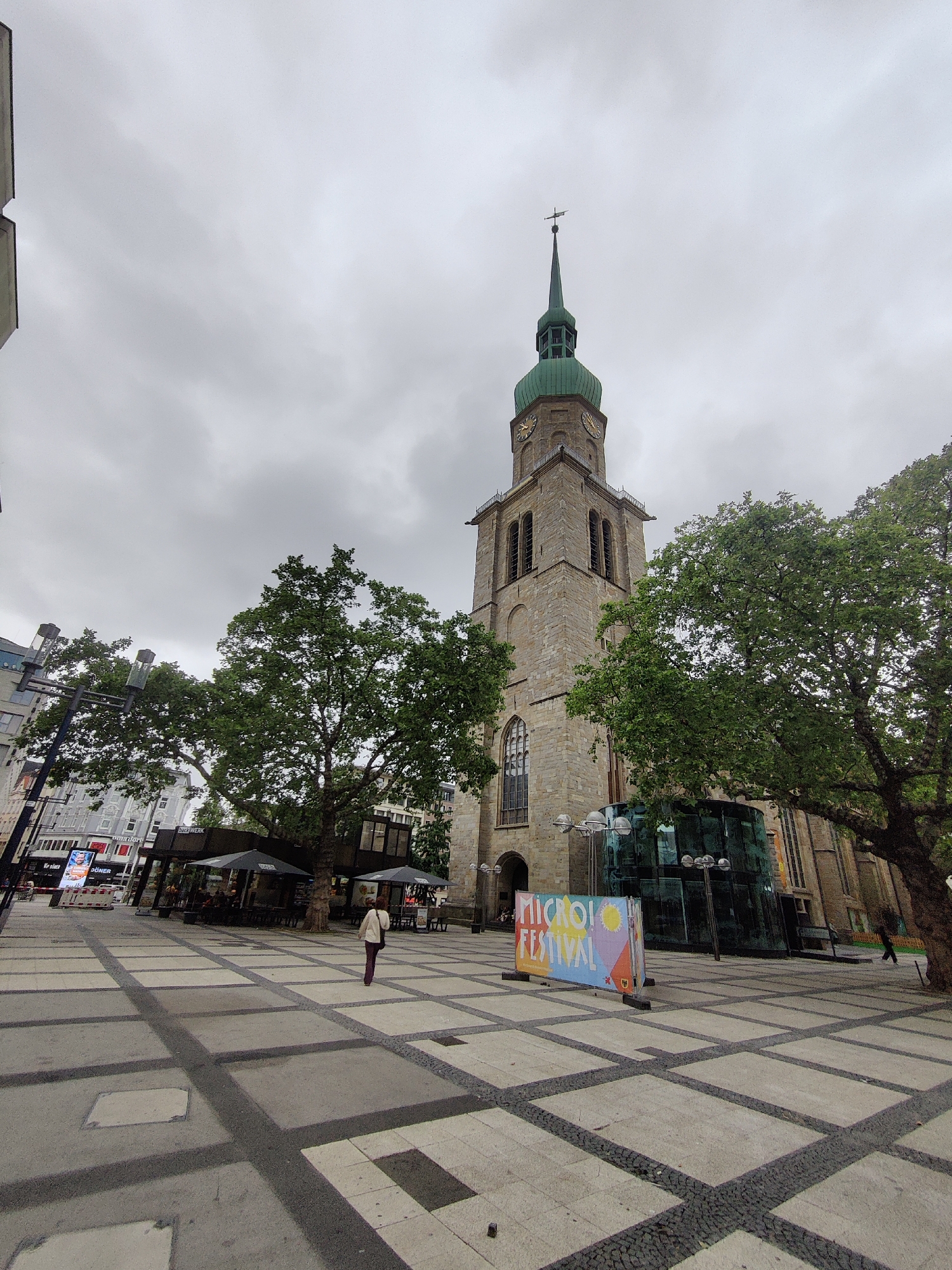 The image depicts a cloudy day in Dortmund, Germany, specifically in front of the Reinoldikirche church. The Reinoldikirche, a prominent landmark in the city, is a stone church with a tall, green-capped spire. In the foreground, a person is walking away from the camera across a paved plaza. The plaza is characterized by a grid pattern of square tiles with dark grout lines. To the right of the church, a modern glass structure is visible, and a colorful banner advertising a "MICRO FESTIVAL" stands in the middle of the plaza. Trees with dense green foliage are scattered throughout the scene, and buildings line the edge of the plaza. Some outdoor seating areas with umbrellas are also present, suggesting a commercial or recreational space. The overall atmosphere is urban and somewhat subdued due to the overcast weather.