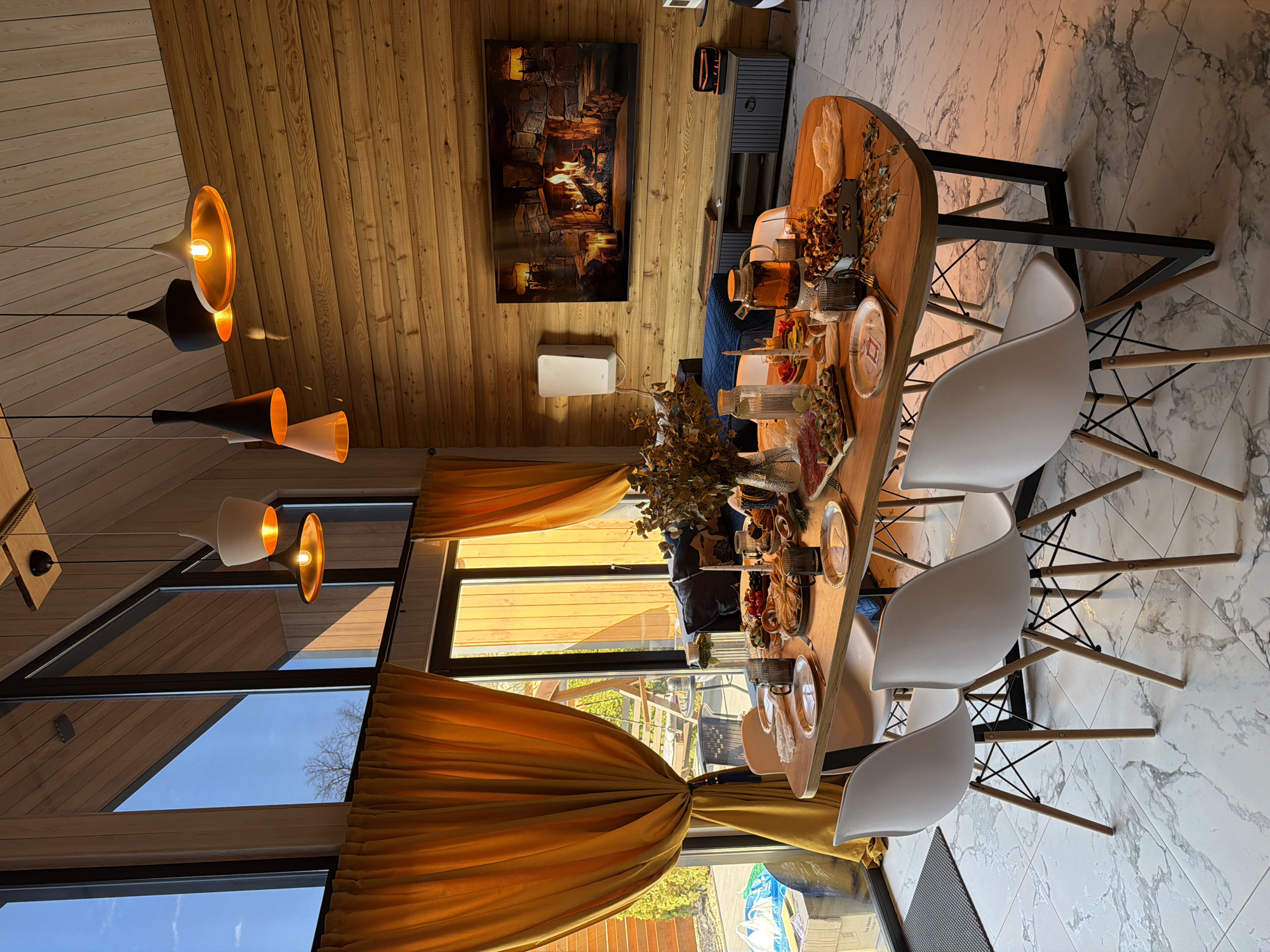The image shows a dining area in a wooden-walled home in Marusino, Russia. The room is bright with light entering through large windows covered with golden drapes. A long, oval wooden table is centrally located, laden with food, drinks, and decorative foliage. Five white plastic chairs with wooden legs are arranged around the table. The floor is covered in light marble tiles with dark grey veining. The ceiling and walls are clad in light brown wood planks. Several modern pendant lights with varying shapes and colors (gold, black, and white) hang from the ceiling. On the wall opposite the windows is a painting of a fireplace above a white box, and a grey cabinet is to the right of the painting. The scene appears to depict a meal setting within a modern wooden home interior.
