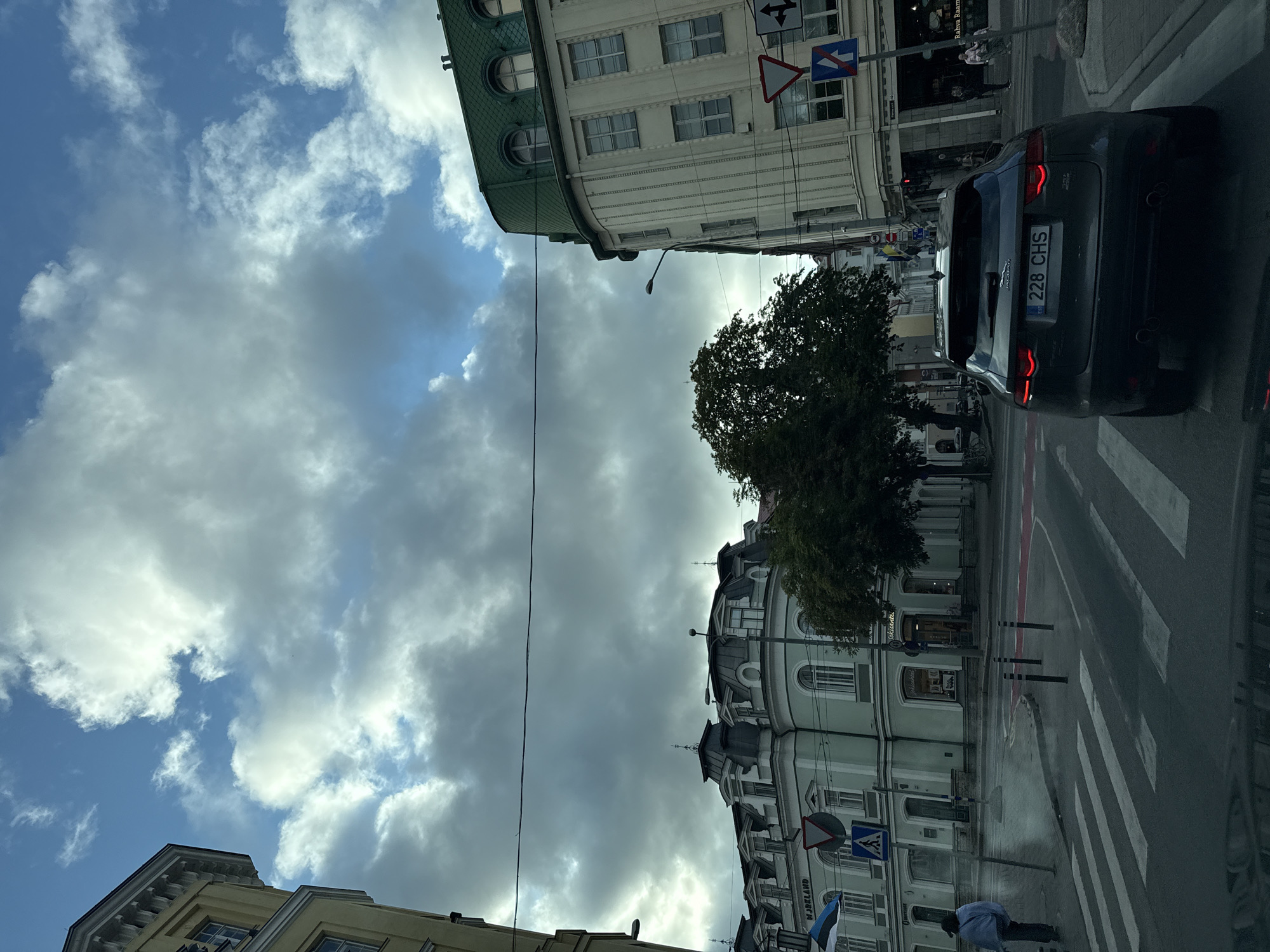 This is a street-level, medium-shot, eye-level, daylight, outdoor, full-color photograph taken from the interior of a vehicle, looking forward. The scene depicts an intersection in Tallinn, Estonia, characterized by overcast skies with patches of blue and white clouds. In the foreground, a dark gray Jaguar F-Pace SUV with license plate "228 CHS" is stopped at a crosswalk. The vehicle's brake lights are illuminated.
The intersection features multiple lanes, crosswalks, and road markings, including red painted sections. Several traffic signs are visible: a triangular yield sign, a blue pedestrian crossing sign, and a sign with a right-turn arrow. Streetlights and utility wires are present.
On the left side of the street, a pale yellow building with architectural details and dormer windows is partially visible. Further down the block, a light green building with a mansard roof and dormer windows displays the word "MJØRKLÄND" in capital letters. A person in a light blue jacket and dark pants is walking away from the camera on the sidewalk to the left of the intersection.
On the right side of the street, a cream-colored building with a distinctive curved facade and a green mansard roof rises several stories. It features multiple windows and has businesses on the ground floor, including one with the sign "Rahva Raamatu". Another person in a pink stroller is being pushed by an unseen individual on the sidewalk to the right. A large, mature tree with dark green foliage stands between the green and cream buildings. The overall impression is a historic European city street scene.