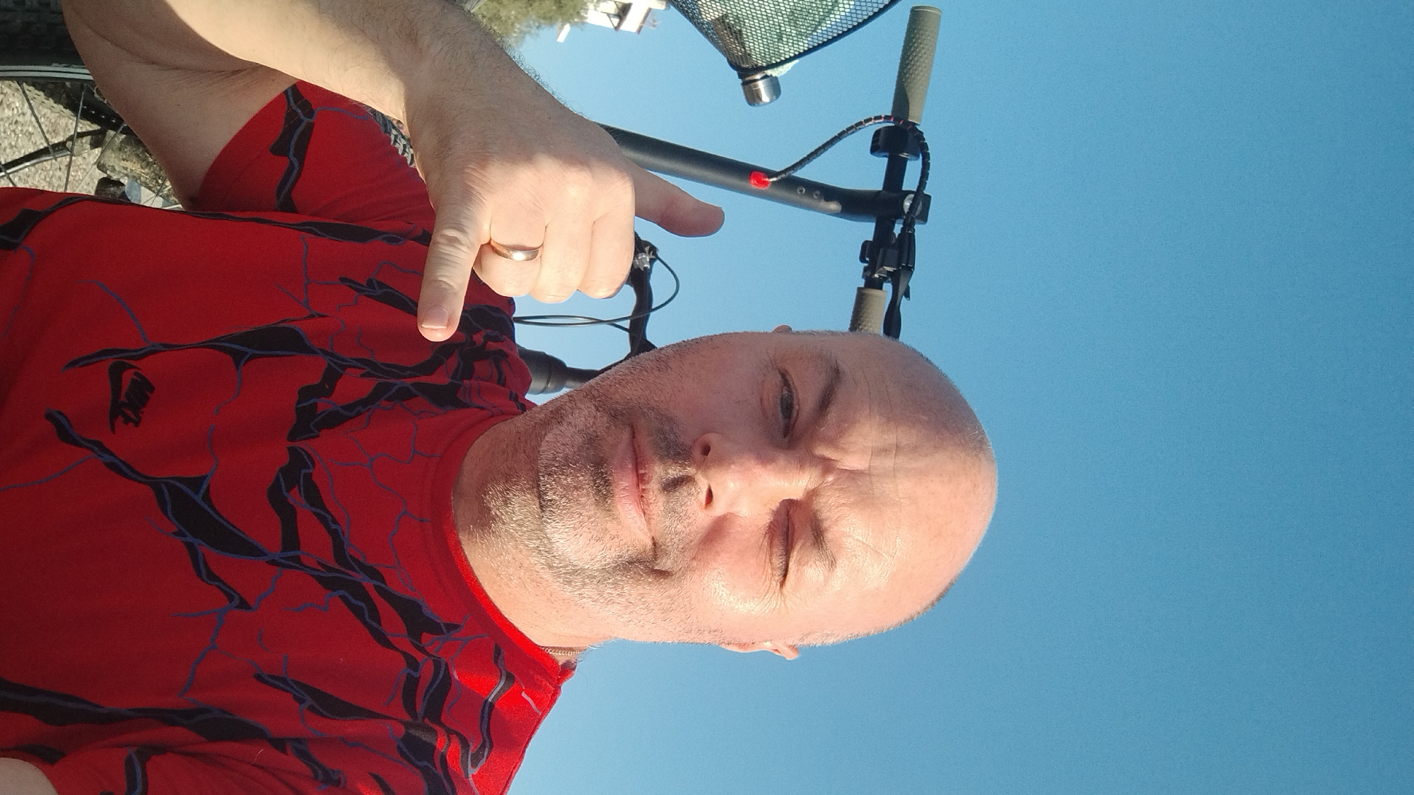 A man is taking a selfie outdoors, with a bright blue sky in the background. He is bald with some stubble and is winking at the camera. He is wearing a red t-shirt with a black and blue web-like pattern and a Nike logo. He is holding up his hand in a "shaka" gesture, with a ring on his left ring finger. Behind him, the handlebars of a black scooter are visible, along with a small basket attached to the front. The image appears to have been taken in the city of Perea, Greece, as indicated by the man's pose, which is a common greeting in that region.