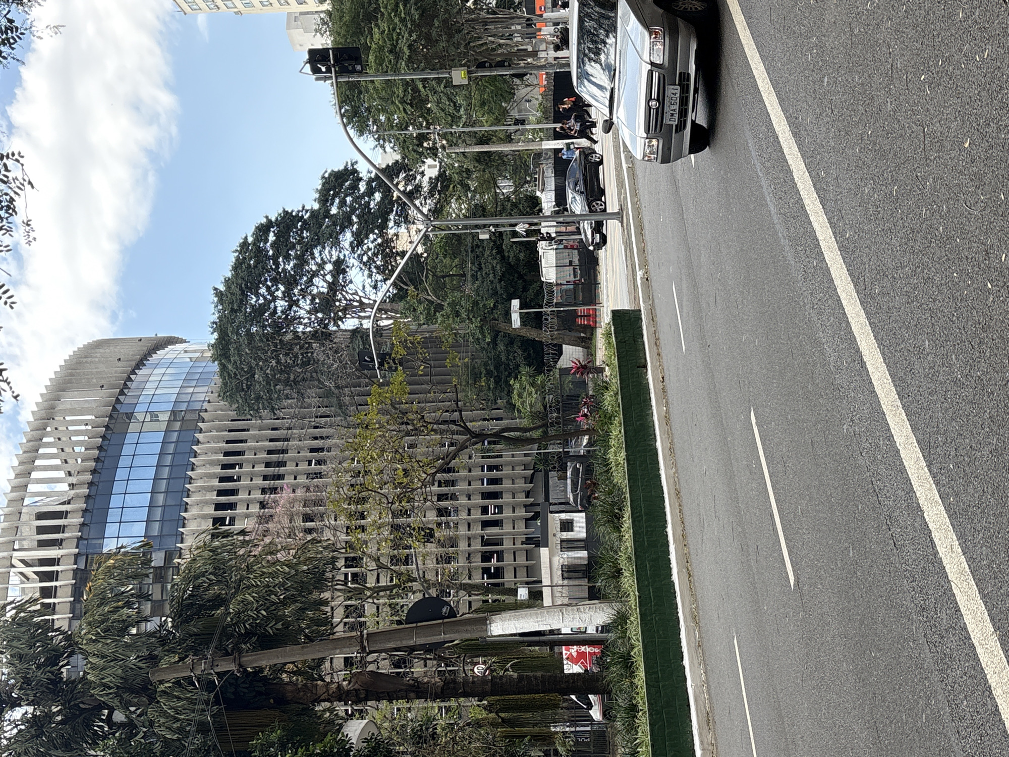 The image captures a street scene in São Paulo, Brazil, specifically on Rua Augusta near the intersection with Rua Haddock Lobo. In the foreground, a silver Fiat Uno with license plate DMA-6041 is driving on the right side of the road. The road itself is paved asphalt with visible white lane markings.
To the left of the road, a green median strip separates the traffic from a sidewalk area. Behind the median, a multi-story concrete building with a distinctive ribbed facade and blue-tinted windows dominates the background. This building appears to be an office or commercial complex. Trees with lush green foliage are interspersed throughout the scene, some reaching high up alongside the building.
Further back, several other vehicles are visible, including a white van and a dark-colored sedan. Pedestrians are also seen walking on the sidewalk in the middle ground. Traffic lights and street lamps are present, indicating an urban environment. The sky above is partly cloudy with a clear blue sky visible. The overall impression is of a busy city street on a bright day.