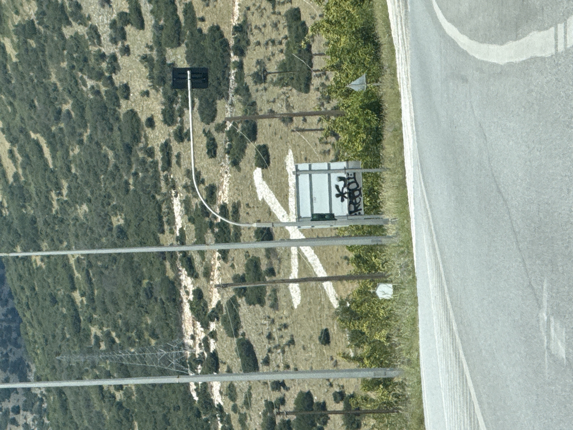 The image depicts a hillside in Prosotsani Municipal Unit, Greece, marked with a large white "X" symbol painted on the terrain. The hillside is covered with sparse green vegetation and some rocky patches. In the foreground, a paved road curves to the right. A traffic light is visible on a pole extending over the road, and a sign with graffiti is situated below it. Several utility poles and wires are also present in the scene.