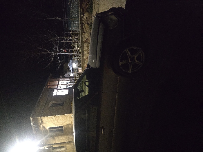 The scene depicts an outdoor nighttime environment in Gorna Mitropolia, Bulgaria. In the foreground, a dark-colored sedan, identified as consistent with a Mercedes-Benz W124 E-Class, is parked on a paved surface. The vehicle's driver's side and front are partially illuminated by an overhead streetlight, revealing multi-spoke alloy wheels. No occupants are visible within the vehicle.
To the left of the vehicle, a two-story building with a light-colored facade, possibly stucco or masonry, is visible. The ground floor of this building features large windows or glass doors that emit interior light. A bright street lamp on a pole stands to the far left, casting significant light on the building and the car's roof.
To the right of the parked vehicle, a low wall topped with a decorative metal fence separates the foreground from a slightly elevated area. Behind this wall and fence, a light-colored gazebo with a dark roof is situated. Several bare deciduous trees are visible in the background, along with a smaller structure featuring a blue roof further beyond. The environment is dark, with artificial light sources providing the primary illumination. No individuals are present in the visible area.