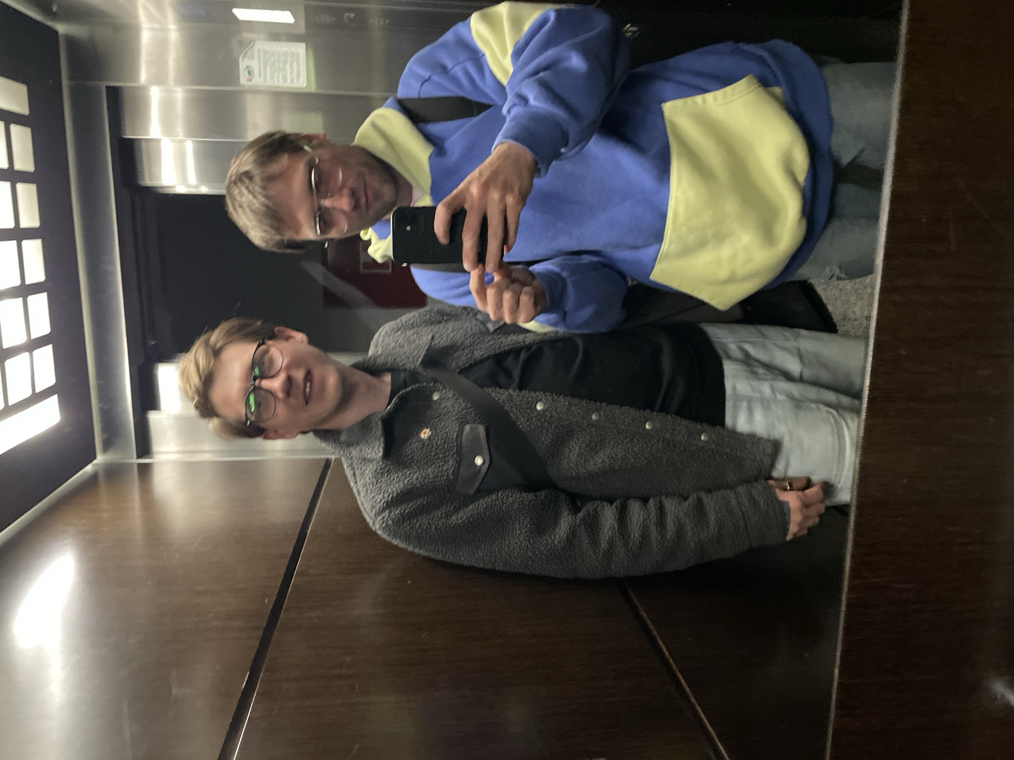 Two men are taking a selfie in an elevator with a mirrored wall. The elevator's interior features a dark wood panel on the lower portion and a stainless steel top half with a vertical row of windows on the left. The man in the foreground wears a gray fleece jacket and glasses. The man behind him wears glasses and a blue and yellow sweatshirt and holds a smartphone. The location is likely not Boreti, Montenegro, as there is no readily available information that places these individuals or this specific elevator within that city.