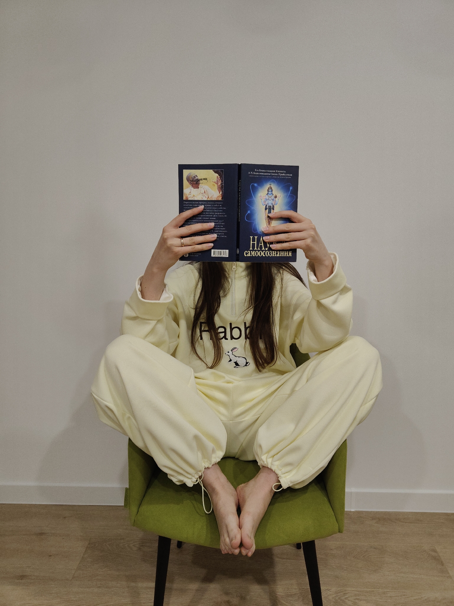 A person is seated cross-legged on a green upholstered chair, holding an open, dark blue book that covers their face. They are wearing a light yellow or cream-colored tracksuit. The top has a partial zipper and features the partially visible word "Rabb" and a white rabbit logo on the chest. The tracksuit bottoms have elasticated cuffs with a drawstring, and the person's bare feet are visible.
The book held by the person has Russian text and imagery. The right cover displays an illustration of a multi-armed deity with a blue aura and the title "НАУКА самоосознания" (The Science of Self-Realization) by "Его Божественная Милость А.Ч. Бхактиведанта Свами Прабхупада" (His Divine Grace A.C. Bhaktivedanta Swami Prabhupada). The left cover features a portrait of an older man in traditional attire, with some Russian text below him, including phrases like "прогресс науки" (progress of science). A barcode is also visible on the left cover. The person's right hand has a ring on the ring finger.
The setting is a simple indoor space with a plain light-colored wall and a wooden floor. The overall impression is one of quiet contemplation or study.