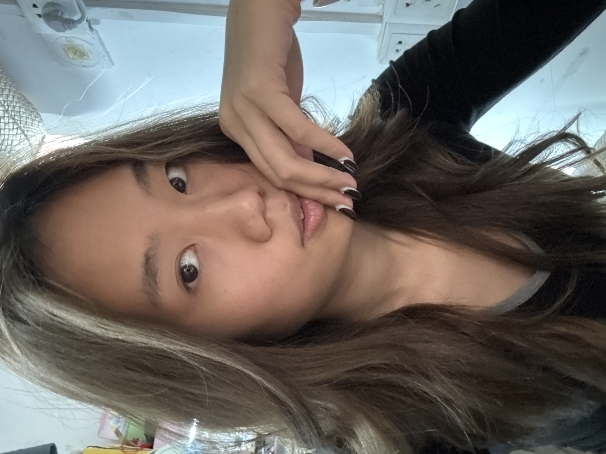 A young woman with light brown hair is pictured in a close-up shot. Her face is partially obscured by her hand, which has a black and white manicure. She is wearing a black long-sleeved shirt. The background is a blurred, light-colored interior with electrical outlets visible. There is no indication of the location of Nan'an District, China.