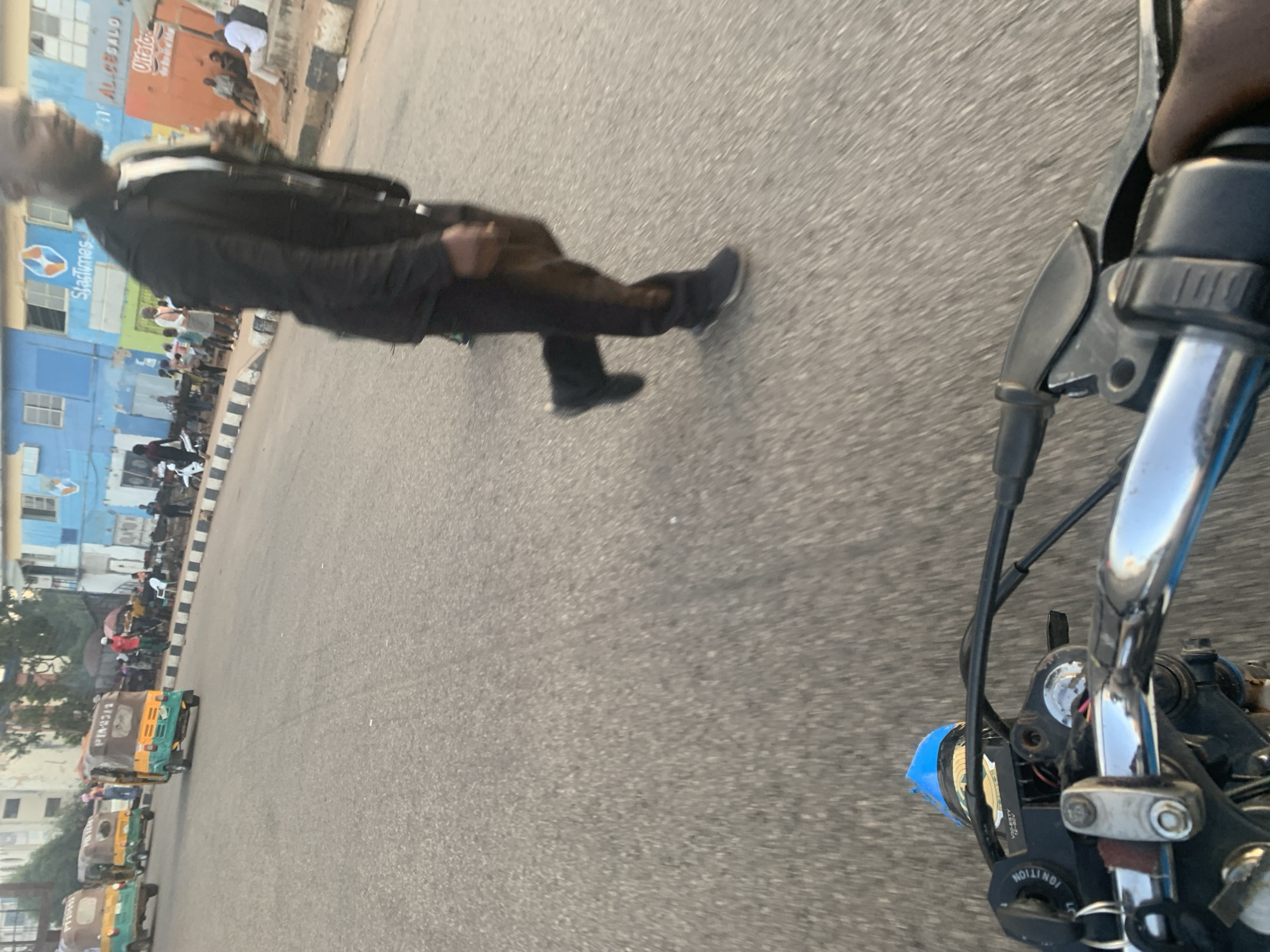 The image captures a street scene in Barakin Lerawi, Nigeria. In the foreground, a first-person perspective from a motorcycle shows the handlebars and part of the dashboard, with a blue object affixed to the top. The road surface is asphalt. To the right, a man in a black jacket and dark pants is walking away from the camera. In the midground, several auto-rickshaws (keke napep) are visible on the road. Further back, pedestrians are present, and buildings with various signage, including "StarTimes" and "ALICE SALO", line the street. The overall environment appears to be an urban area with moderate activity.