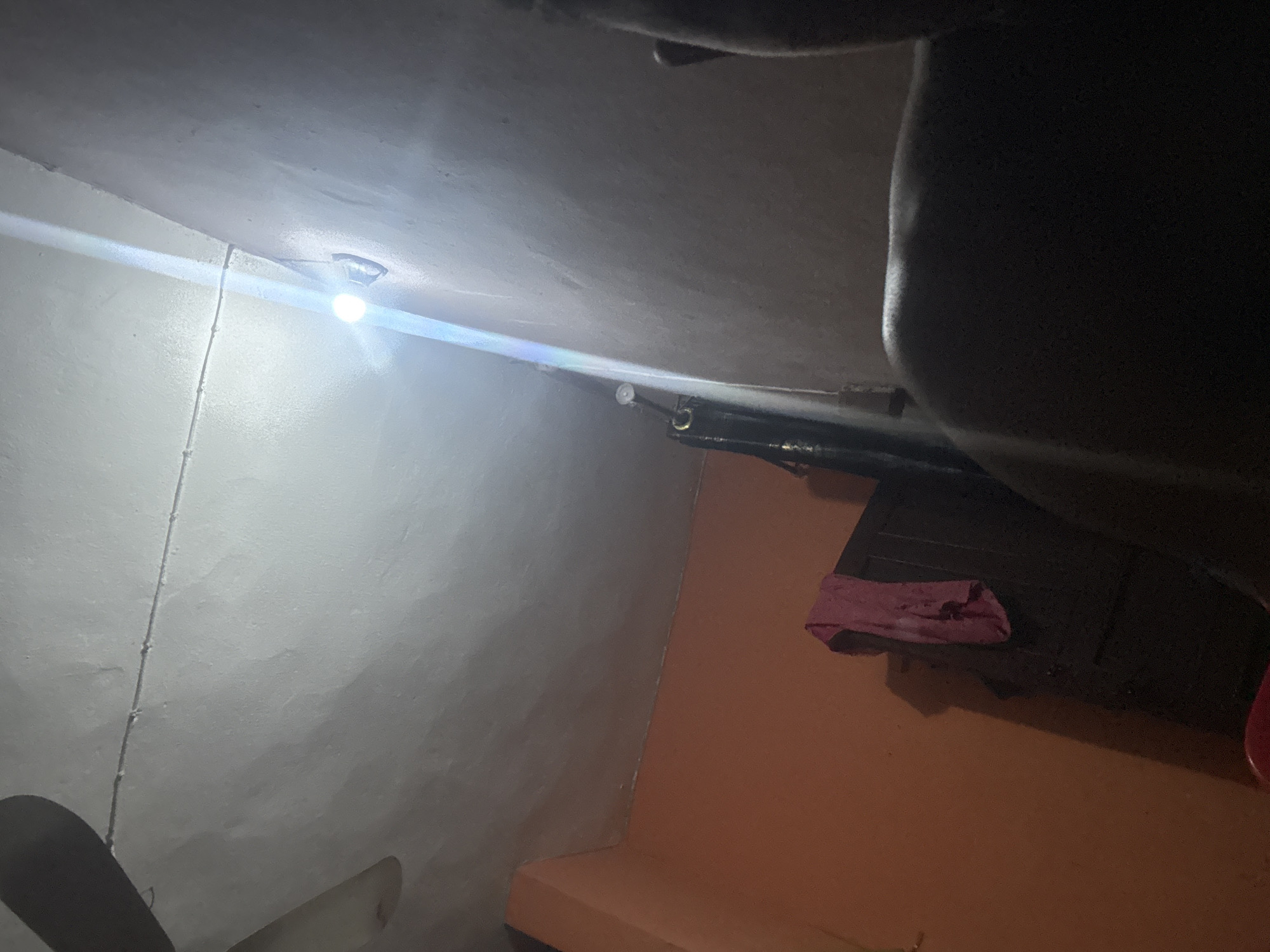 The image depicts an indoor scene, likely a bedroom, in Onitsha, Nigeria. A ceiling fan is visible in the upper left corner, partially illuminated by a bright light bulb hanging from a wire attached to the ceiling. The ceiling appears to be textured and painted a light color, possibly white or off-white. A portion of an orange-colored wall is visible on the left side of the image. To the right of the orange wall, a dark wooden cabinet or wardrobe is present, with a pink piece of fabric draped over its top. In the foreground, on the right side, a person's back and shoulder are visible, suggesting they are sitting or leaning against something. The lighting is harsh, with the light bulb casting strong glare and creating deep shadows. The overall impression is of a modest, likely residential space in Onitsha.