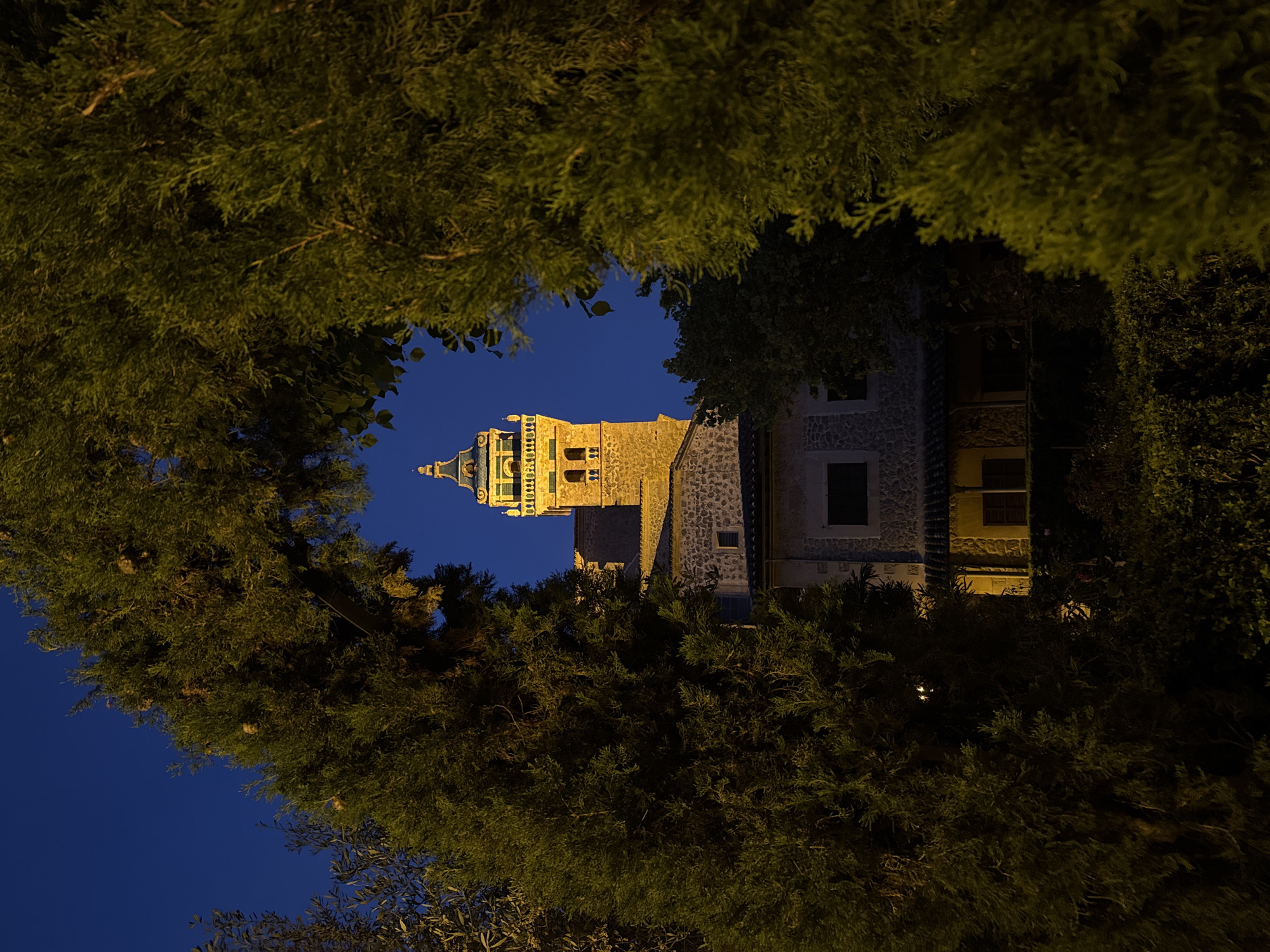 The image depicts a tower framed by foliage at night in Valldemossa, Spain. The tower is illuminated, showing a light stone structure with green and gold accents on the top. A portion of a building with windows is visible next to the tower. The sky is a dark blue. The surrounding trees create a natural frame around the tower.