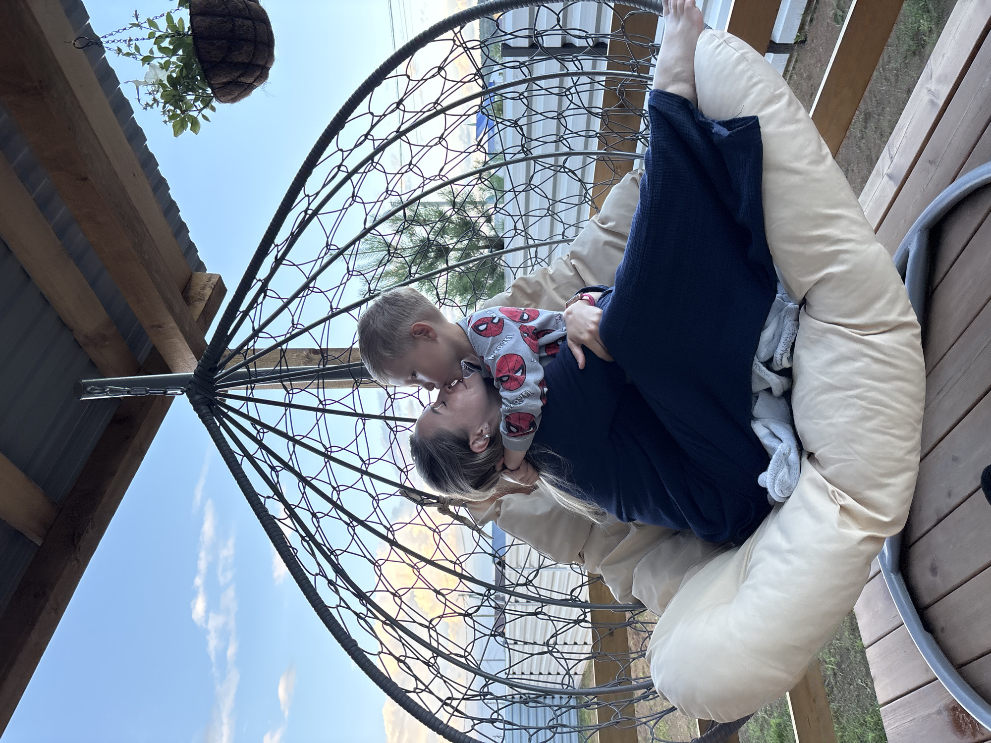 In this photograph, a mother and her young son are sharing a tender moment while seated in a large, woven hanging chair. The mother is wearing a dark blue robe or jumpsuit, and her son is dressed in pajamas featuring a Spider-Man pattern. They are positioned closely, with the boy leaning in to kiss the side of his mother's face. The hanging chair is supported by a dark metal frame and filled with light-colored cushions, creating a cozy seating area.
The scene appears to be outdoors, possibly on a porch or balcony, given the wooden deck visible in the background and the presence of a railing. Above and to the left, part of a wooden roof structure with corrugated metal roofing can be seen, along with a hanging basket containing greenery and white flowers. The sky is a clear blue with a few wispy white clouds, suggesting a pleasant daytime setting. The lighting indicates it's likely late afternoon or early evening, with the sun casting soft light. The overall mood is one of warmth, affection, and relaxation. There is no visible text in the image.