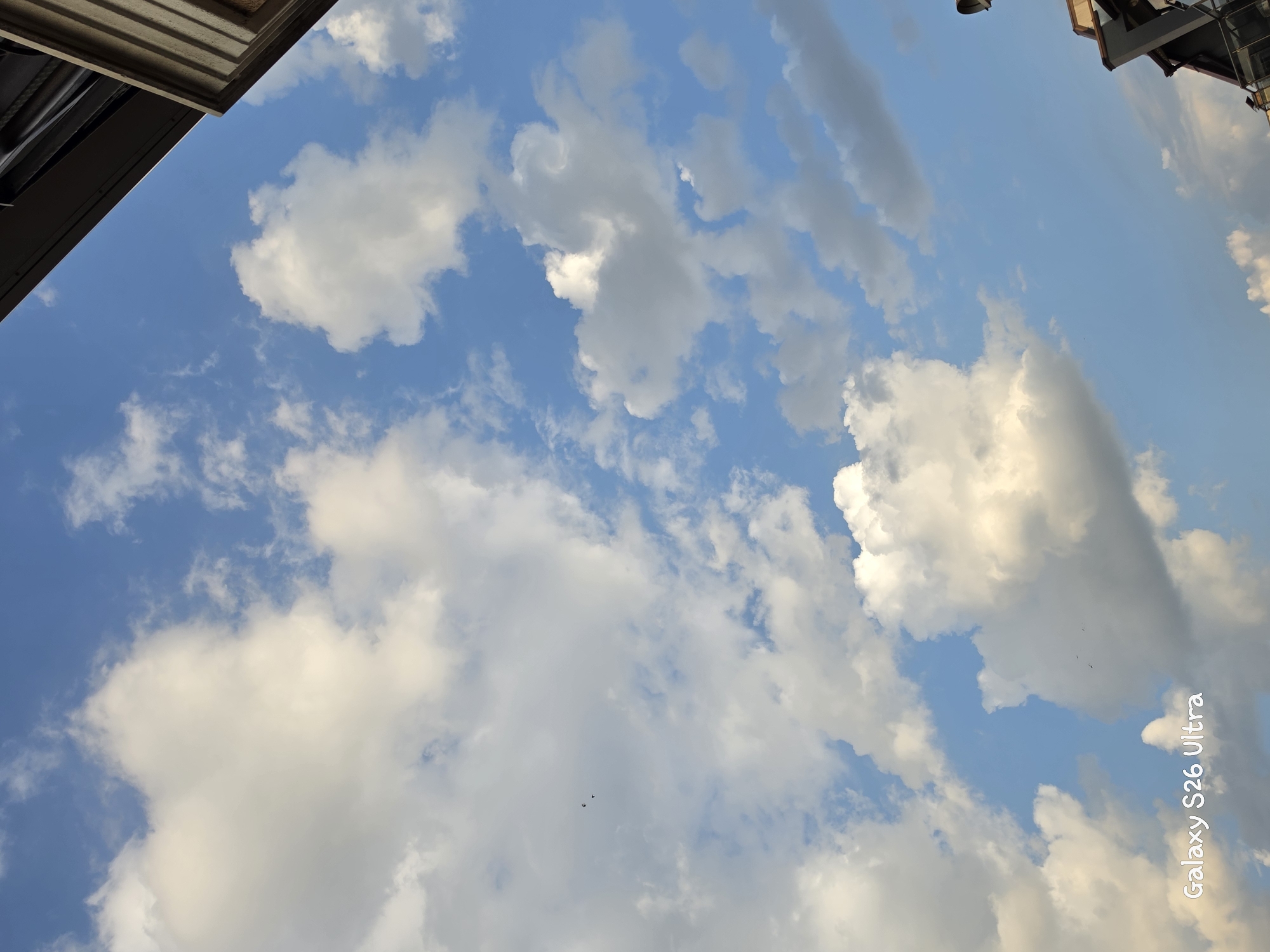 This is a low-angle, vertical photograph of a bright blue sky filled with fluffy white and grey clouds. In the upper left and upper right corners of the image, parts of buildings or structures are visible, suggesting an urban or semi-urban setting looking upwards. The lighting indicates it is daytime, likely late afternoon or early evening, given the warm tones in some of the clouds and the long shadows possibly cast by the unseen structures. The sky is dynamic, with scattered cumulus clouds and some streaky cirrus clouds. Two small dark shapes, likely birds in flight, are visible in the lower center of the image. The overall mood is serene and expansive.
Visible text: "Galaxy S26 Ultra" is written vertically in white on the right side of the image.