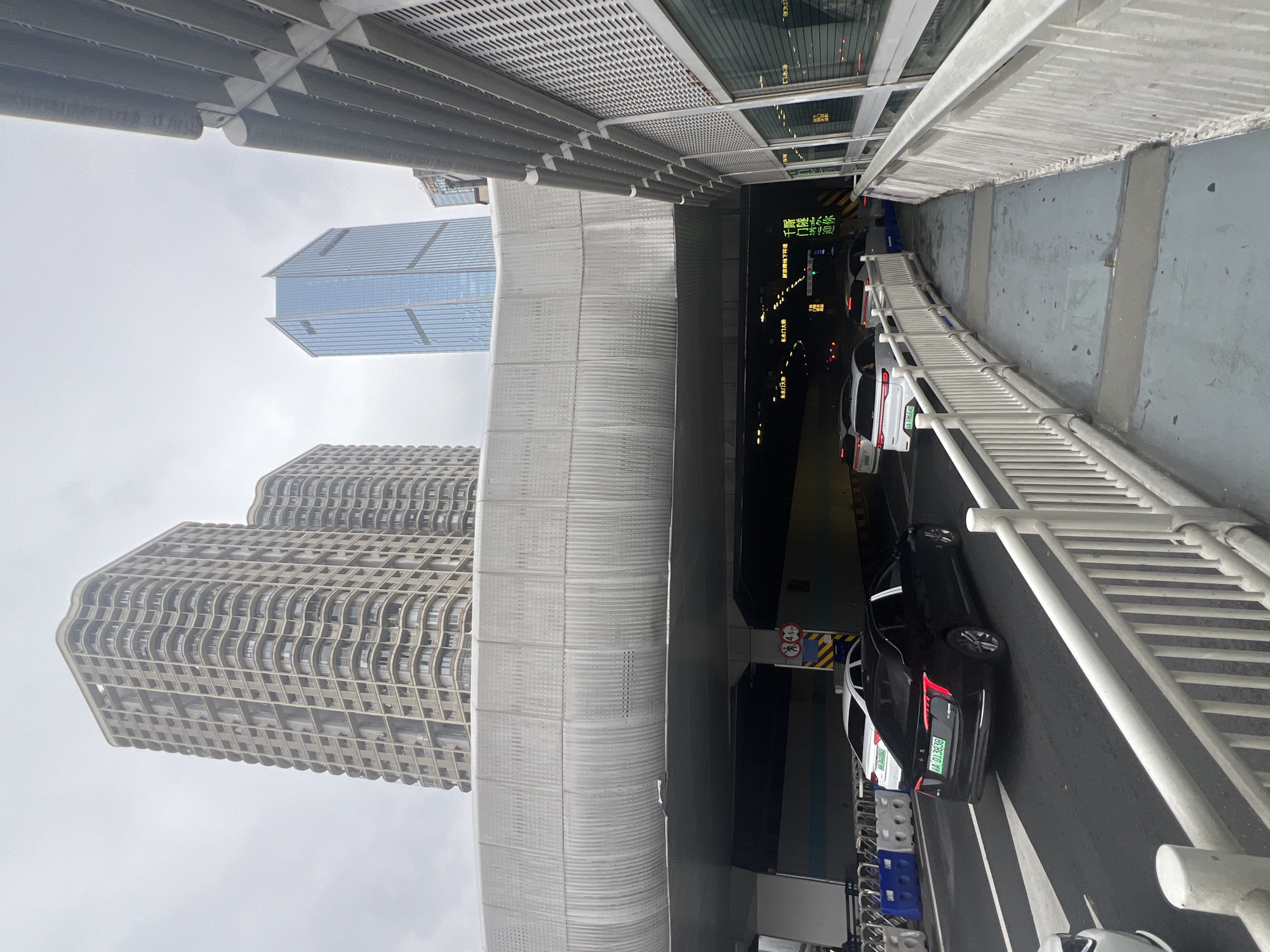 This is a daytime, eye-level, outdoor, wide shot of a road leading into a tunnel under a large overpass structure. The tunnel entrance is visible with signs and lights. Several cars are on the road, some entering the tunnel. The road is flanked by white railings and concrete barriers. In the background, tall modern buildings, including a glass skyscraper, are visible against an overcast sky. The architecture suggests an urban environment in Yuzhong District, China.