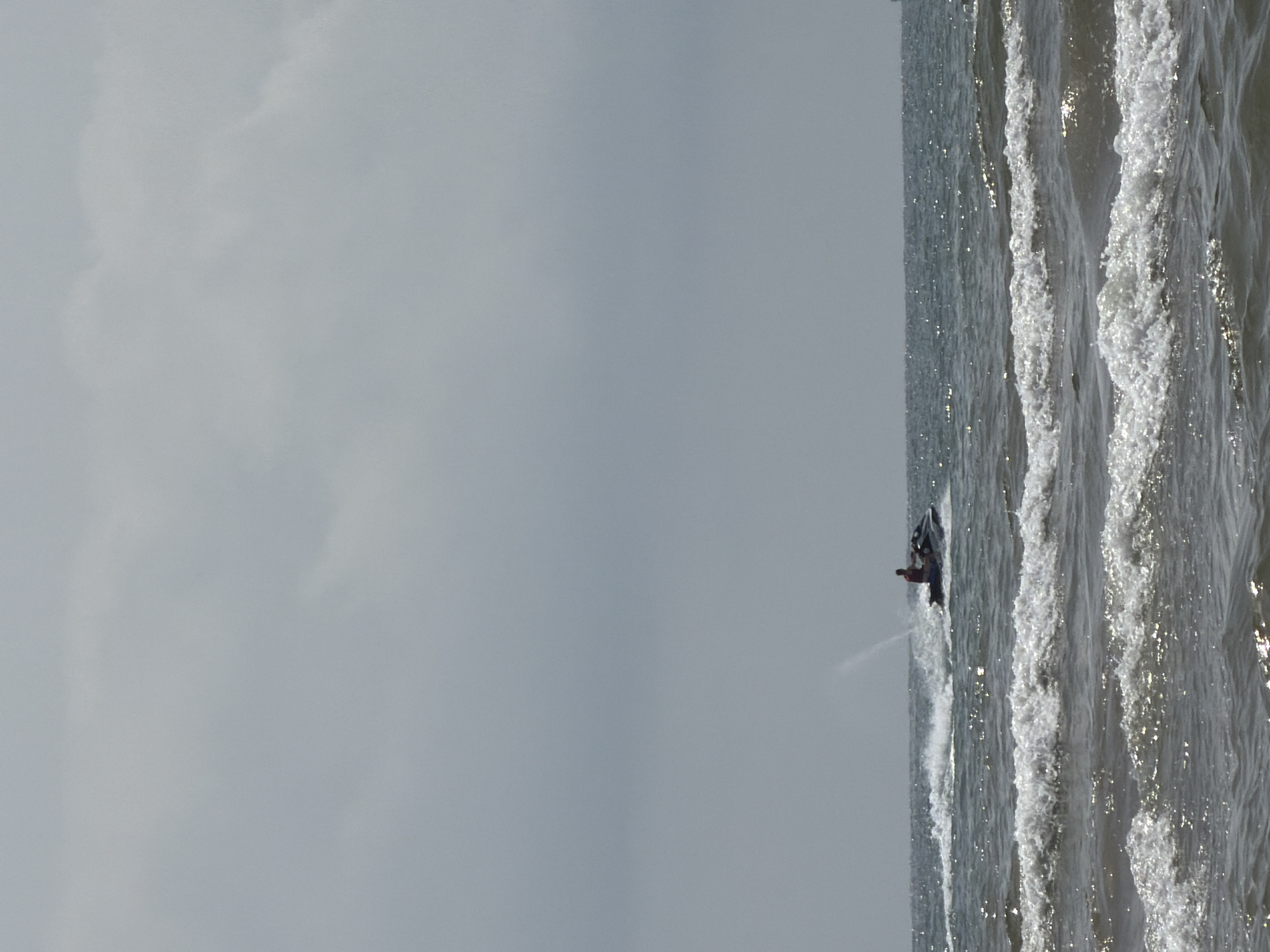 An overcast sky hangs above a body of water with moderate waves. A person on a jet ski is visible in the middle distance, riding across the water and creating a spray. The jet ski is dark in color, and the person is wearing a red shirt. The water is choppy, with white foam cresting on the waves. The scene appears to be at a beach or coastal area, though no specific landmarks are visible to identify the city or country. The overall impression is one of a somewhat gray and active day at the seaside.