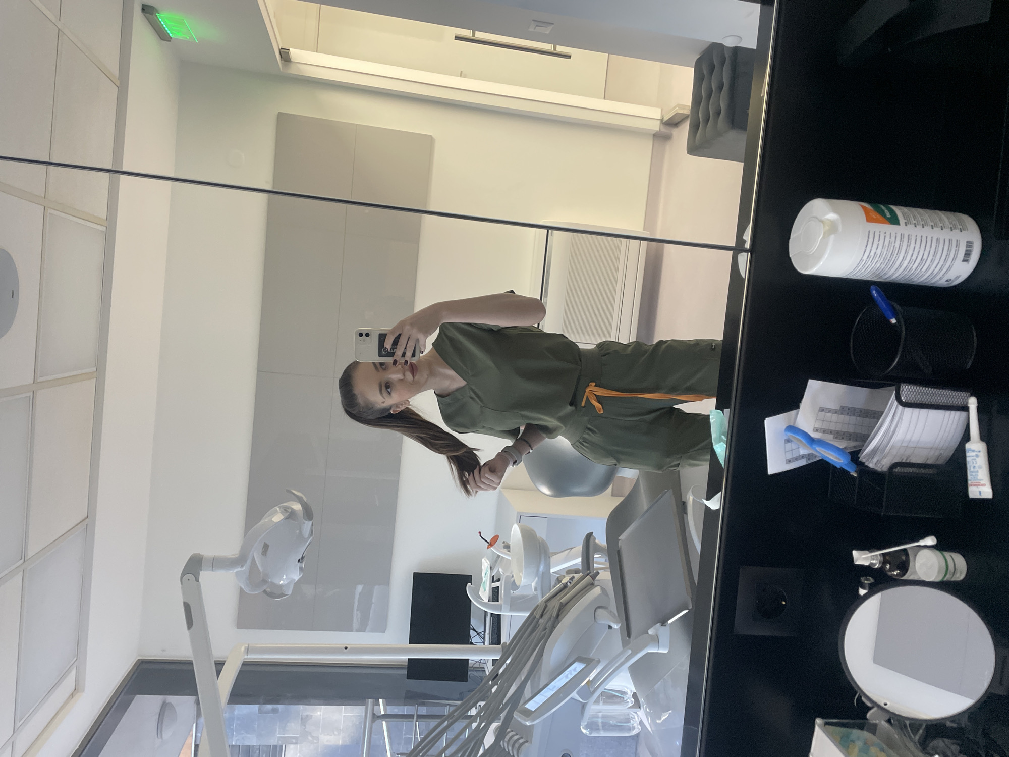 The image shows the reflection in a mirror of a woman in olive green scrubs with orange drawstring, holding a smartphone in her right hand. She has long brown hair pulled back in a ponytail.
The reflection shows a modern dental clinic. There is a dental chair and associated equipment, including a dental light. A monitor is visible in the background. The walls are white with light gray paneling.
The immediate foreground shows the side of a black cabinet containing various items. These include a white cylindrical container with an orange label, a black wire mesh container with pens and paper, a small container with blue handles, a mirror, cotton swabs, and other small items.
There is a green exit sign on the ceiling. The location is likely a dental clinic in the city of Sofia, Bulgaria.