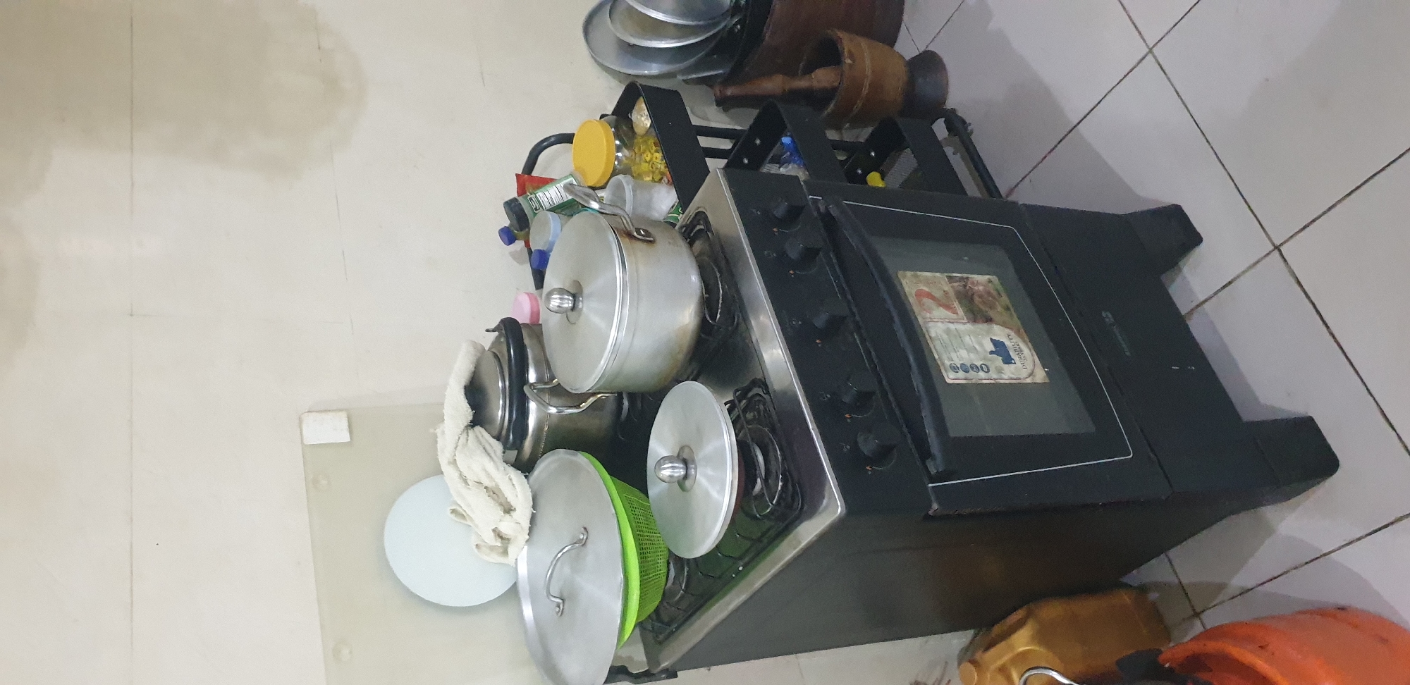 The image depicts an indoor kitchen area, located in Barden Yana, Nigeria. The central focus is a black freestanding gas range with an integrated oven, positioned on a tiled floor.
The stove's cooktop features four burners. On the left rear burner, a large silver-colored pot with a matching lid rests on a green plastic colander. On the left front burner, a smaller silver-colored pot with a domed lid is present. The right front burner is empty. On the right rear burner, a medium-sized silver-colored pot with a lid is positioned. Directly behind the large pot on the left, a silver metallic thermos or kettle, topped with a white towel, stands.
To the left of the stove, on the tiled floor, a yellow plastic jerrycan is visible, with an orange container (possibly another jerrycan or a gas cylinder) partially visible further left.
To the right of the stove, a black, two-tiered rolling utility cart is situated. The top shelf holds several bottles and jars; one clear jar has a yellow lid and contains small yellow items, while another holds a white substance. A dark green bottle and other assorted containers are also on this shelf. The bottom shelf is less clear but appears to hold additional items. Adjacent to the cart on the floor, a wooden mortar and pestle are placed. A stack of metallic plates or pot lids leans against a dark wooden drum or container to the right of the mortar.
The walls are light-colored, possibly off-white, and exhibit subtle horizontal lines, suggesting paneling or plaster divisions. Some faint, irregular discolorations are visible on the upper right portion of the wall. Above the stove, a light blue circular chopping board is propped against the wall, and a small, rectangular white object is adhered to the wall segment to its left. The floor consists of light-colored, square ceramic tiles with dark grout lines. The oven door on the stove features a partially visible label with text including "DEURABILITY." No individuals are present in the scene.