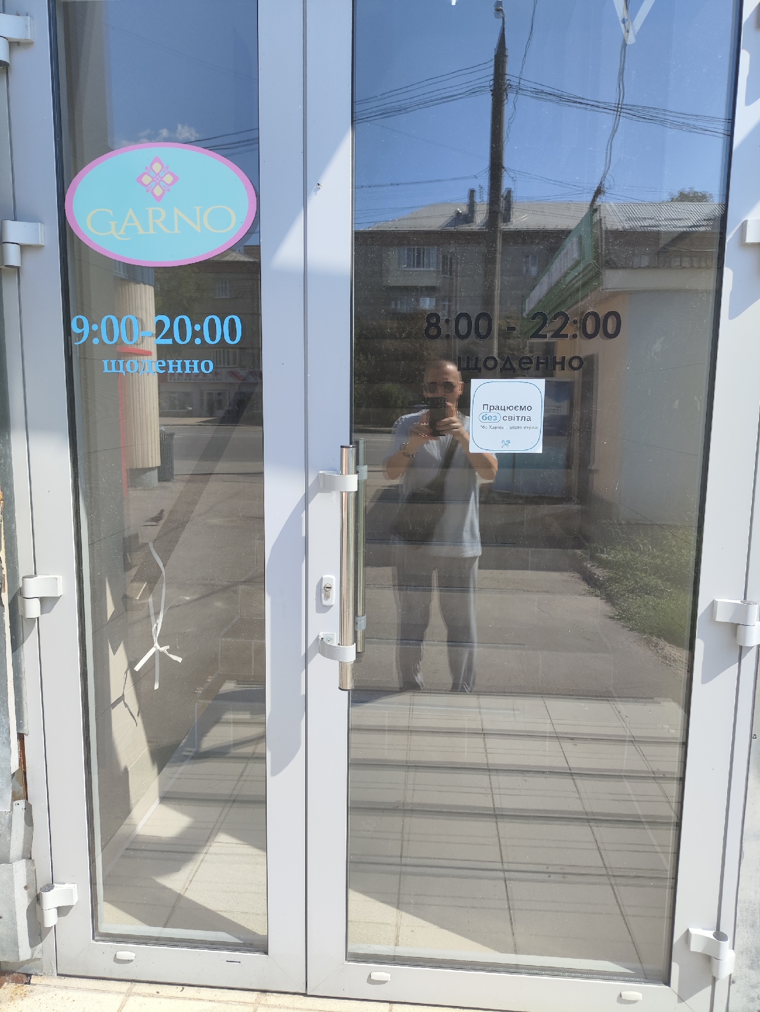 The image shows the glass doors of a store with signage. On the left door, a pink and blue oval logo with the text "GARNO" is visible. Below the logo, blue text reads "9:00-20:00" and "щоденно" (daily). On the right door, black text reads "8:00 - 22:00" and "щоденно". A white sticker with blue text is also visible on the right door, saying "Працюємо (без) світла" (We work (without) light) and at the bottom, "*Без Харкова - місто-героїв" (*Without Kharkiv - city of heroes). A person wearing a grey t-shirt and grey pants, holding a smartphone, is reflected in the glass, taking a picture of their reflection. The person is standing on a tiled sidewalk outside the store. The background shows buildings and power lines. This scene is likely in Kharkiv, Ukraine, as indicated by the text on the sticker.