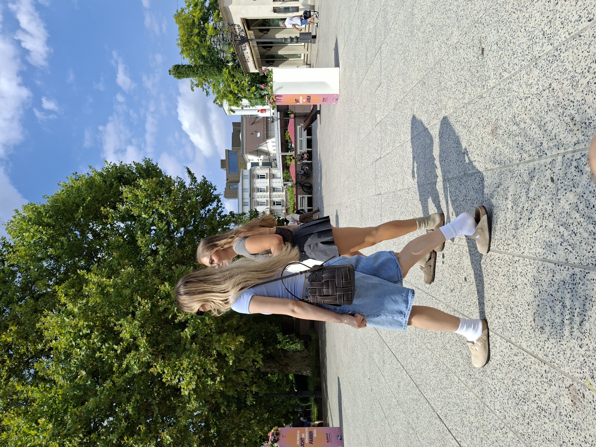 Two young women walk across a paved plaza in Baden-Baden, Germany, on a sunny day. The woman in the foreground, on the left, has long blonde hair and wears a light blue t-shirt, denim shorts, white socks, and tan clogs. She carries a dark brown woven handbag. The woman behind her has long blonde hair and wears a grey t-shirt and a dark grey pleated skirt, also with tan clogs. They are walking away from the camera. In the background, a large tree with dense green foliage dominates the left side of the frame. Beyond the tree, buildings with various architectural styles are visible, including a multi-story building with a modern glass facade. To the right, a building with "PHOTO R//KÖNIG" signage is partially visible. Several outdoor seating areas with red umbrellas are set up in front of some establishments. A white, rectangular display stand with colorful graphics stands in the plaza. A street lamp with multiple fixtures is mounted on a pole near the building with the "PHOTO R//KÖNIG" sign. The sky is blue with scattered white clouds. The ground is paved with light-colored, speckled tiles.