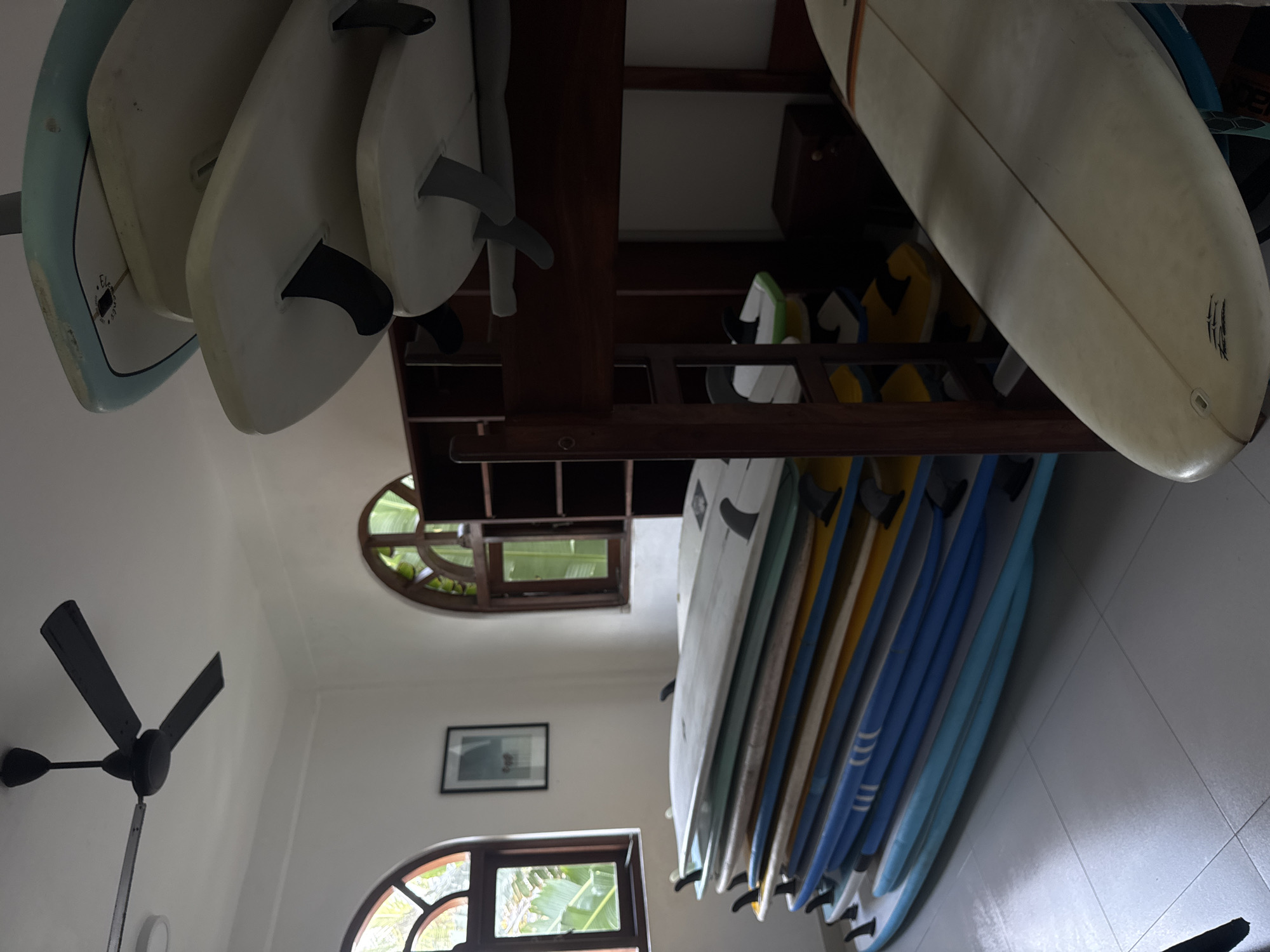 The scene depicts the interior of a building in Goviyapana, Sri Lanka, specifically a room where surfboards are stored. The room is painted white and has a tiled floor. A ceiling fan with three blades is visible in the lower left corner. There are two arched windows, one near the ceiling and one near the floor, both allowing natural light to enter. A framed picture hangs on the wall between the two windows. The primary focus is on a rack of surfboards. Some surfboards are stacked horizontally on the top shelf of the rack, while others are stored vertically in the rack below. The surfboards are of varying colors, including white, yellow, and blue.