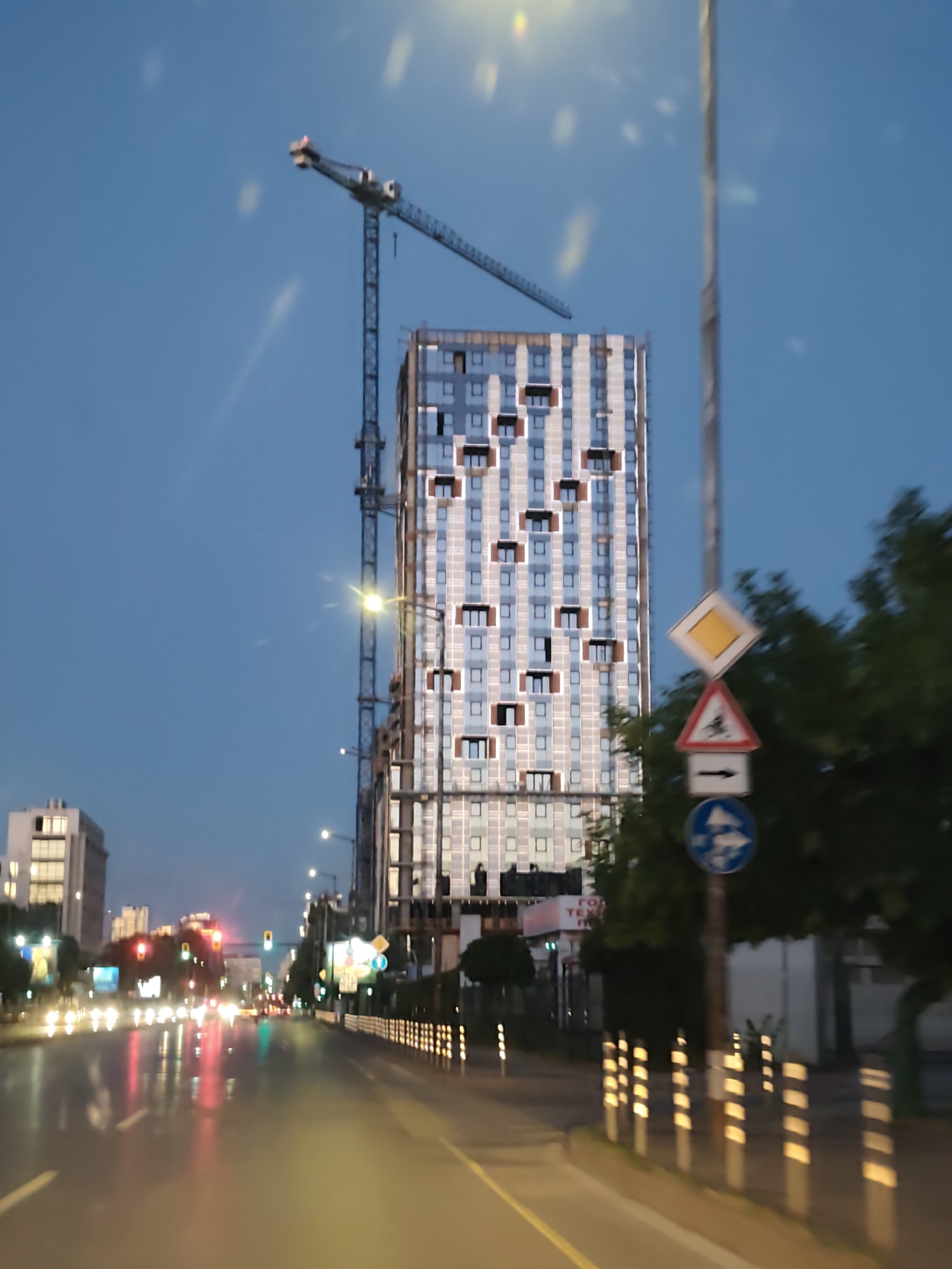 The image depicts a street scene in Sofia, Bulgaria, at dusk or twilight. The road is wet, reflecting car headlights and streetlights. On the right side of the road, there's a sidewalk with trees and street signs. A tall building is under construction, with a crane visible alongside it. The building has a distinctive pattern of windows, with some balconies. In the background, other buildings and traffic lights are visible, blurring into the distance. The road is marked with yellow lines and reflective posts.