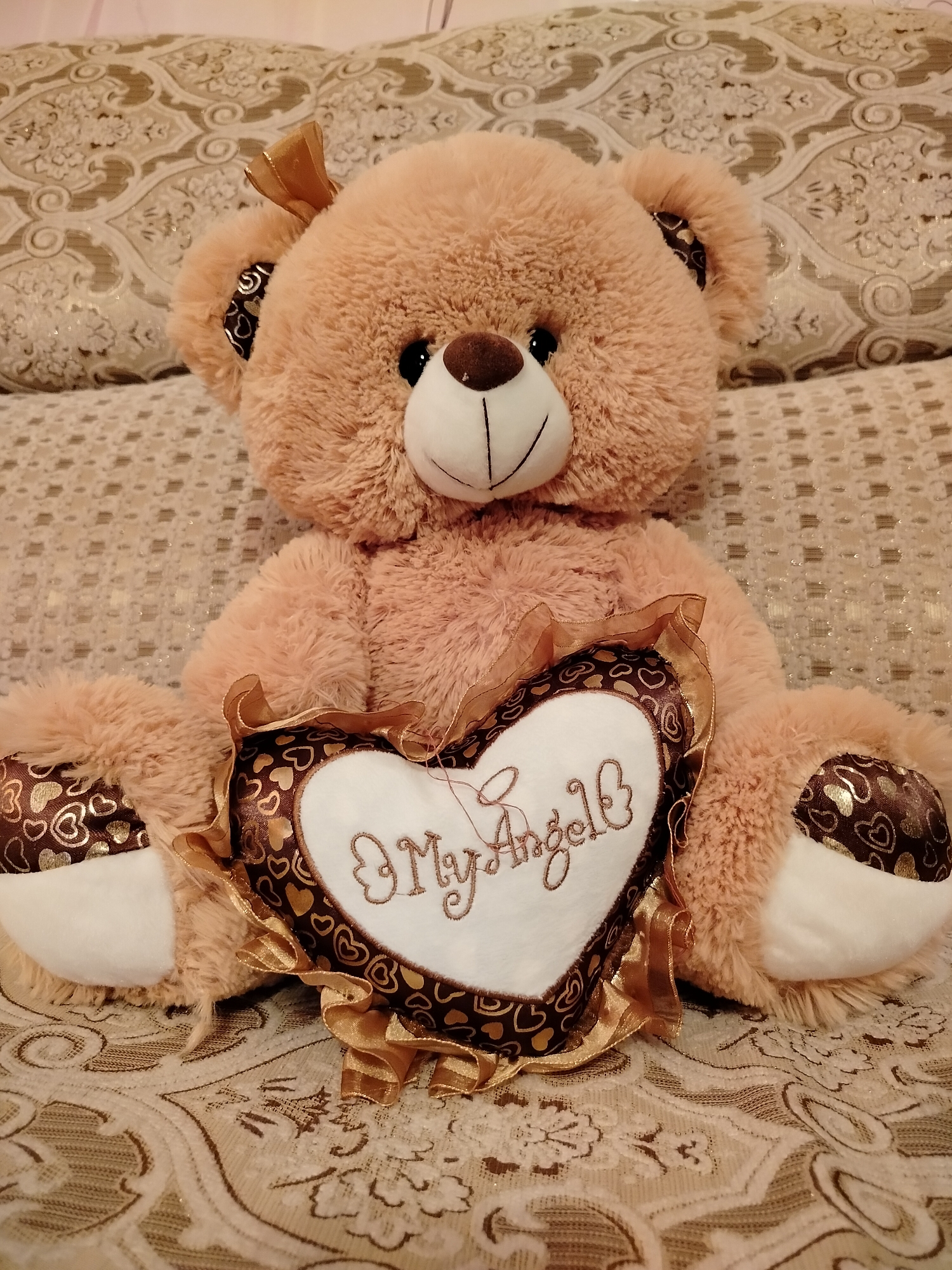 A fluffy, light brown teddy bear is seated on a textured, patterned surface, likely a sofa or bed, with pillows behind it. The teddy bear is holding a heart-shaped pillow with the embroidered words "My Angel" in cursive. The heart pillow has a brown border with gold heart patterns and a ruffled gold ribbon trim. The teddy bear has a stitched smile and dark button eyes. The background features pillows and fabric with intricate, possibly damask-style, patterns in neutral tones. The image does not provide any information about the location in Chișcăreni, Moldova.
