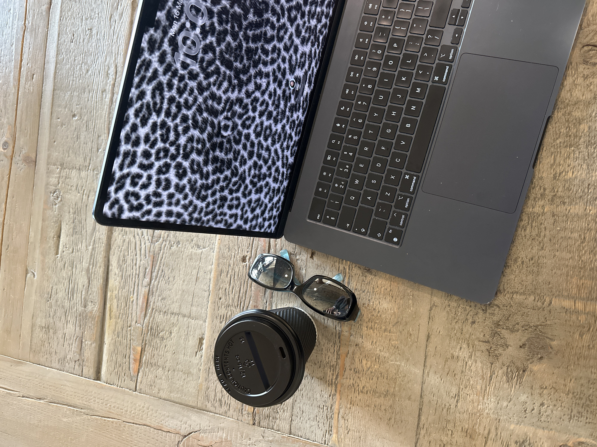 The scene displays a collection of objects arranged on a light-toned wooden surface, consistent with a table or desk, located in the city of London, United Kingdom. An open, dark grey laptop occupies the right side of the frame. Its screen exhibits a black and white leopard print wallpaper, overlaid with a digital clock displaying "10:00" and the date "Mon 16 Mar" in the upper right quadrant, and a small user icon labeled "Kitty" at the bottom center. To the upper left of the laptop, a black disposable coffee cup with a ribbed exterior and a black lid is positioned. The lid is marked with text including "CAUTION CONTENTS HOT" and "12/16 oz". Between the coffee cup and the laptop's keyboard, a pair of black-framed glasses featuring prominent light blue temple tips rests on the wooden surface. The wooden surface itself shows distinct grain patterns and minor textural variations. No individuals are visible within the frame.