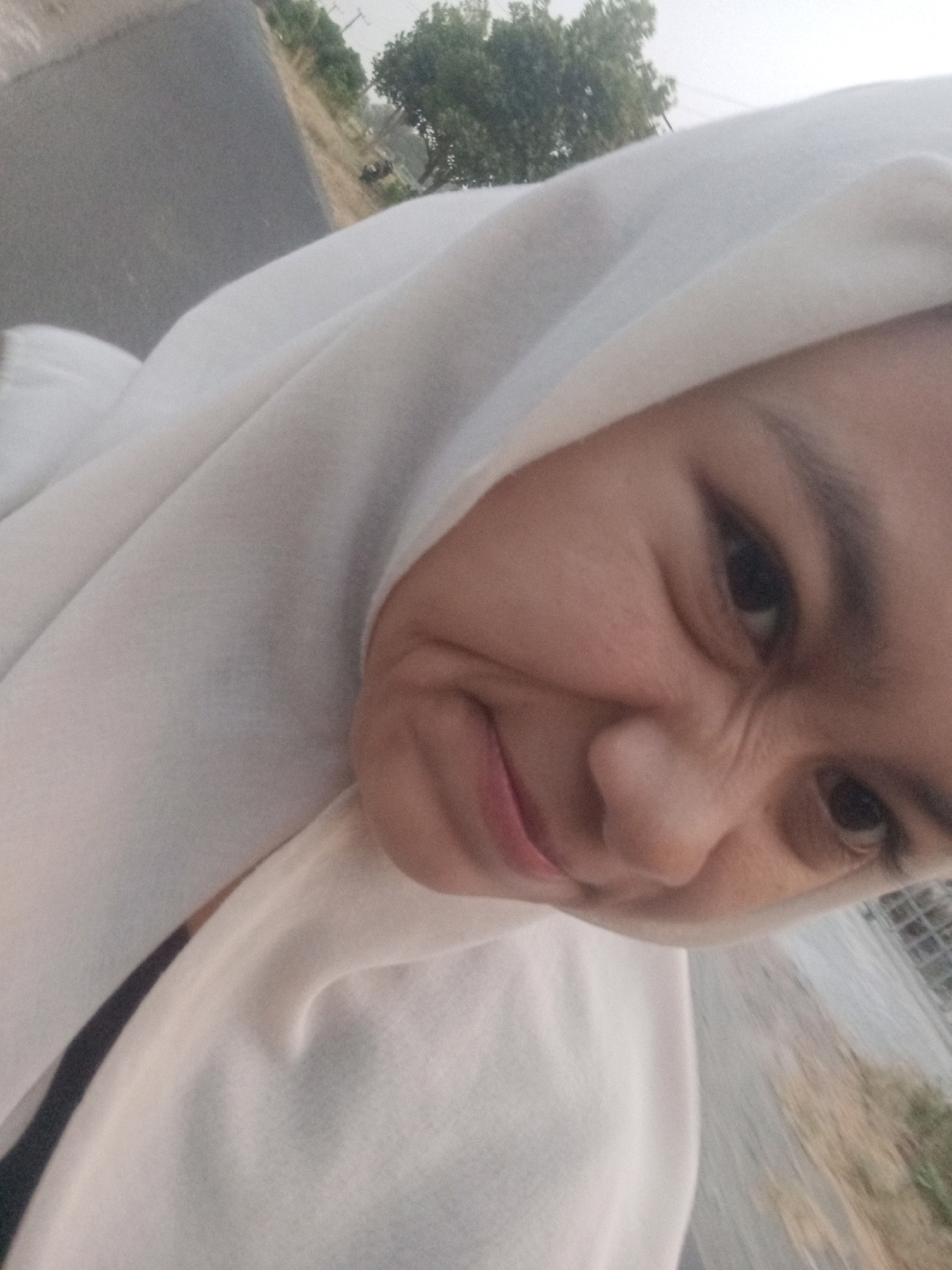 A close-up, low-angle, over-the-shoulder selfie captures a person wearing a white hijab, with their face contorted into a playful grimace. The individual's eyes are narrowed, and their mouth is pursed, with the tip of their tongue visible. The background suggests an outdoor setting, possibly a roadside in Plemahan, Indonesia, indicated by the presence of a gray asphalt road, dry vegetation, and some trees. A faint glimpse of a fence or metal structure is also visible. The overall lighting is dim, characteristic of an overcast sky or late evening.