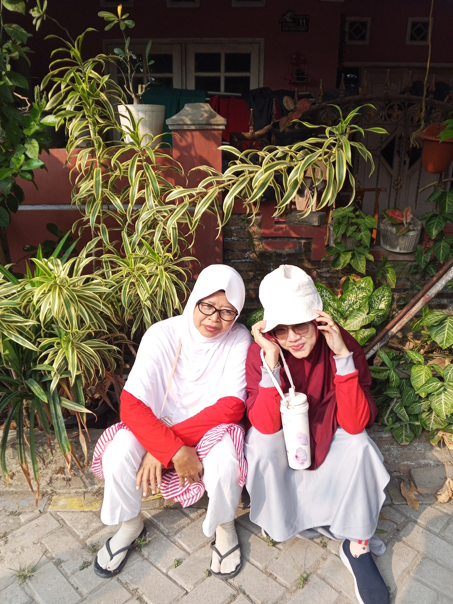 Two women, dressed in Muslim attire, are seated outdoors in what appears to be a residential area in Ciruas, Indonesia. The woman on the left wears a white hijab, glasses, a white long-sleeved top with red accents, and white pants. She also has a red and white striped cloth draped over her lap and wears black flip-flops. The woman on the right wears a white bucket hat, sunglasses, a maroon hijab and long-sleeved top with red accents, and a light gray skirt. She holds a white cylindrical water bottle with a floral pattern. They are positioned in front of lush green plants and a reddish-brown building. The ground is paved with interlocking bricks. The scene suggests a casual outdoor setting, likely in the afternoon given the lighting.