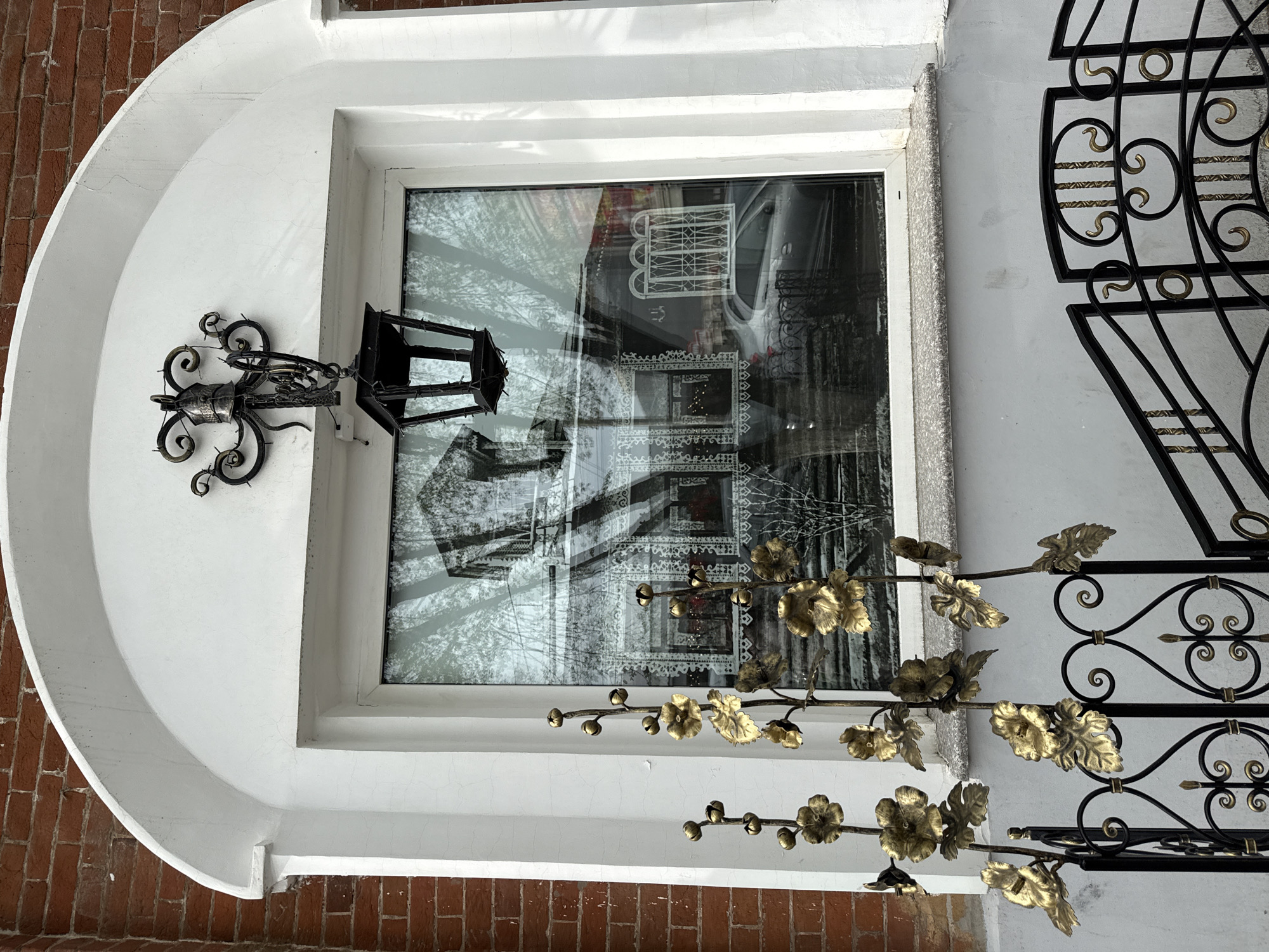 The image shows a window of a building in Votkinsk, Russia. The window is white-framed and has a rounded arch above it. A wrought-iron lantern is attached to the wall to the left of the window. The window reflects the image of another building across the street, and in front of the window, partially obscuring the view, is a wrought-iron decoration featuring golden flowers and leaves. To the right of the window is an ornate wrought-iron fence. The wall surrounding the window is painted white. Brick is visible beneath the window and to the left of the arch.