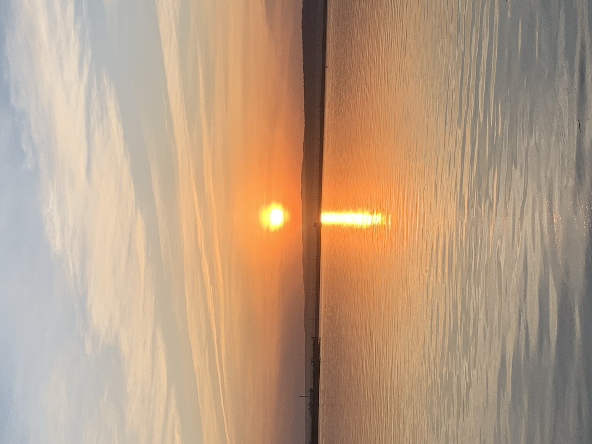 The image depicts a sunset over water, likely a body of water in Chaikovsky, Russia. A bright orange sun is positioned on the horizon, its reflection creating a shimmering path on the water's surface. The sky transitions from pale blue with clouds on one side to orange and yellow hues near the horizon. A dark, thin strip of land is visible on the horizon line, separating the sky from the water. The water surface is rippled, reflecting the colors of the sky and sun. No people are visible in the image. The scene is serene and focuses on the natural beauty of the sunset.