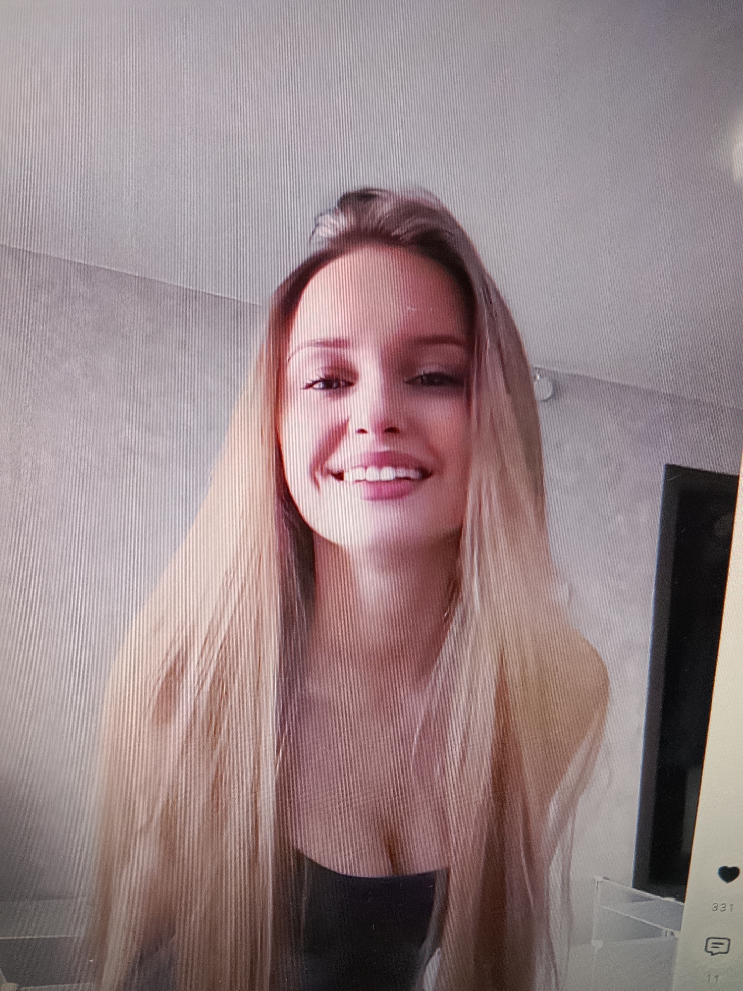 A young woman with long, blonde hair is looking at the camera and smiling. She is wearing a black top. The background appears to be a wall with a textured pattern, possibly in a room. There are some faint lines visible on the image, which may indicate a screen or a projection. To the right, a dark vertical object is visible, and a small portion of a white object can be seen below it. Social media icons and numbers are also visible in the lower right corner of the image. The overall lighting is soft, with the woman's face being well-lit.
