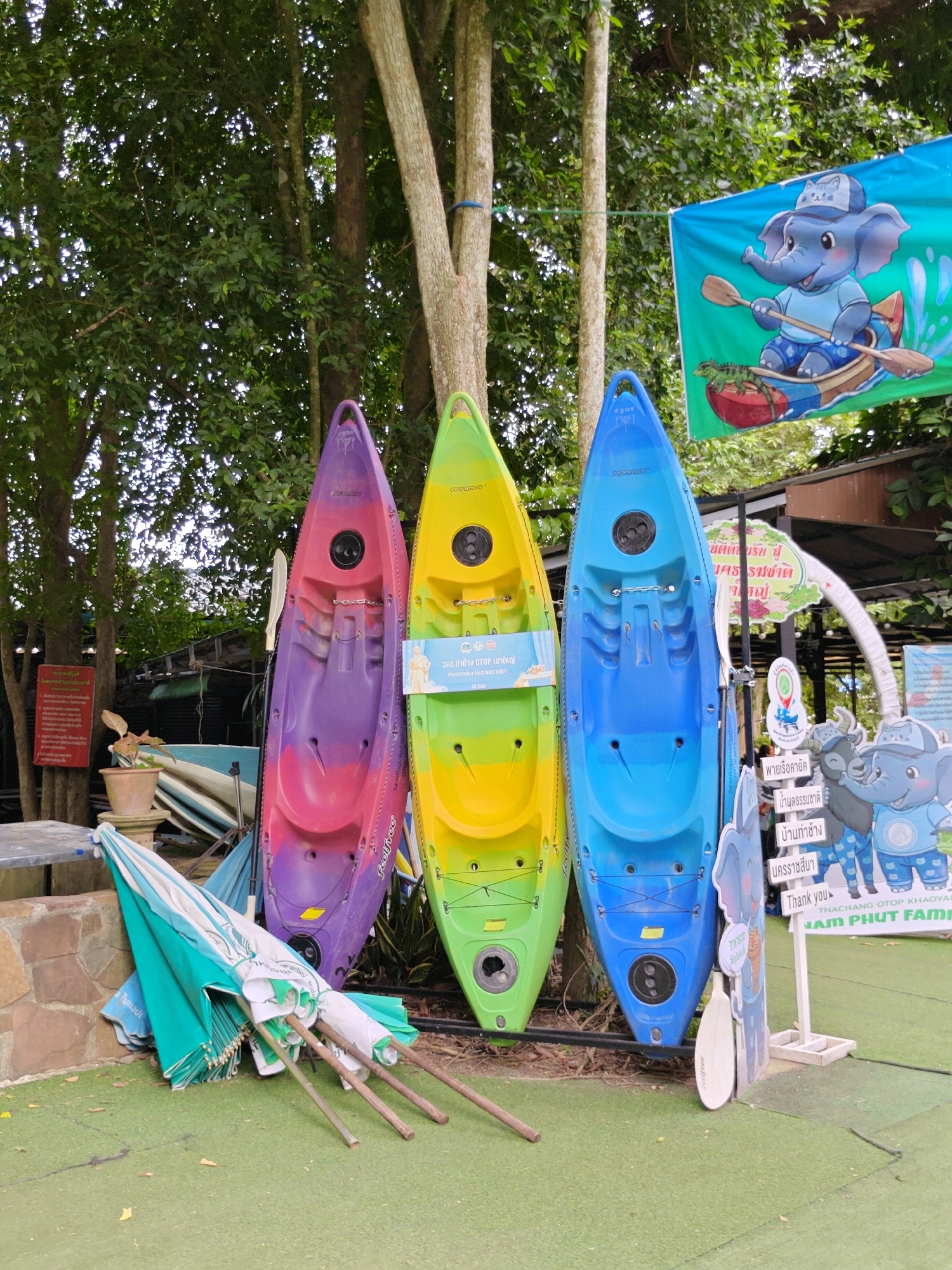The image depicts an outdoor scene in Tambon Mu Si, Thailand, featuring three kayaks standing upright against trees. The kayaks are colored purple and pink, yellow and green, and blue. To the left of the kayaks, a pile of closed green and white striped umbrellas rests on the ground. In the background, lush green foliage from trees forms a canopy overhead. A colorful banner with an illustration of a cartoon elephant in a boat is hung on the right side. Various signs with Thai writing and cartoon elephants are also visible. The ground is covered in a green artificial turf.