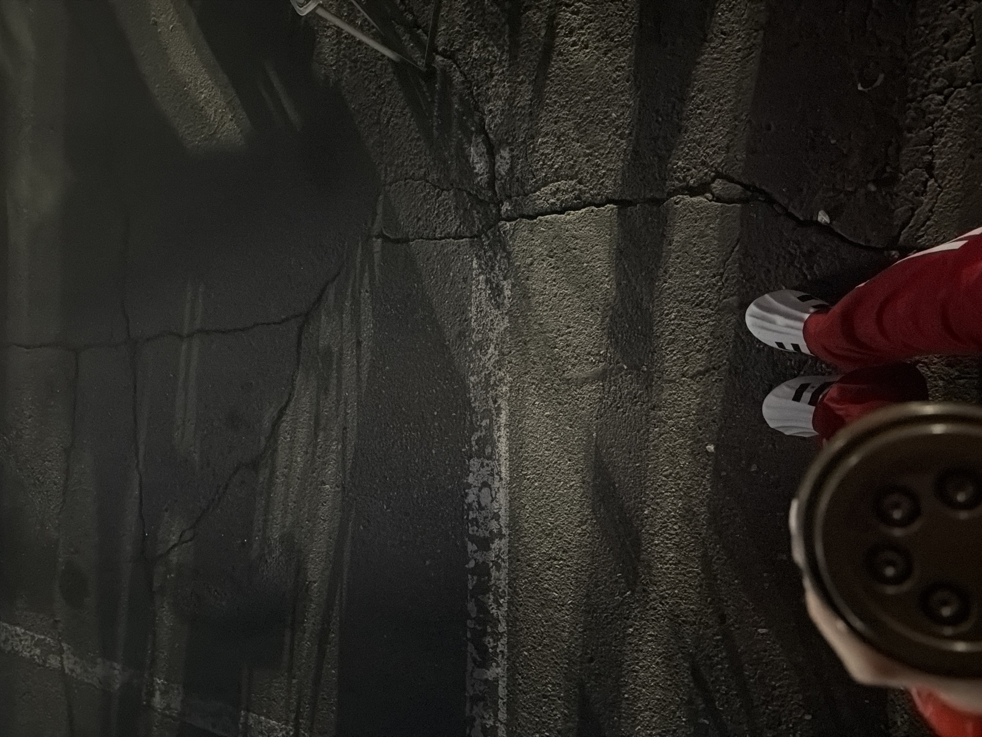 The image shows a section of cracked asphalt ground with a person standing on it. The person is wearing white sneakers with black stripes and red pants. A dark, circular object with four visible holes is held in the person's hand. The ground is textured with cracks and uneven surfaces. The scene appears to be dimly lit, possibly at night. The location within the city of Unknown, Unknown is not discernible from the image.