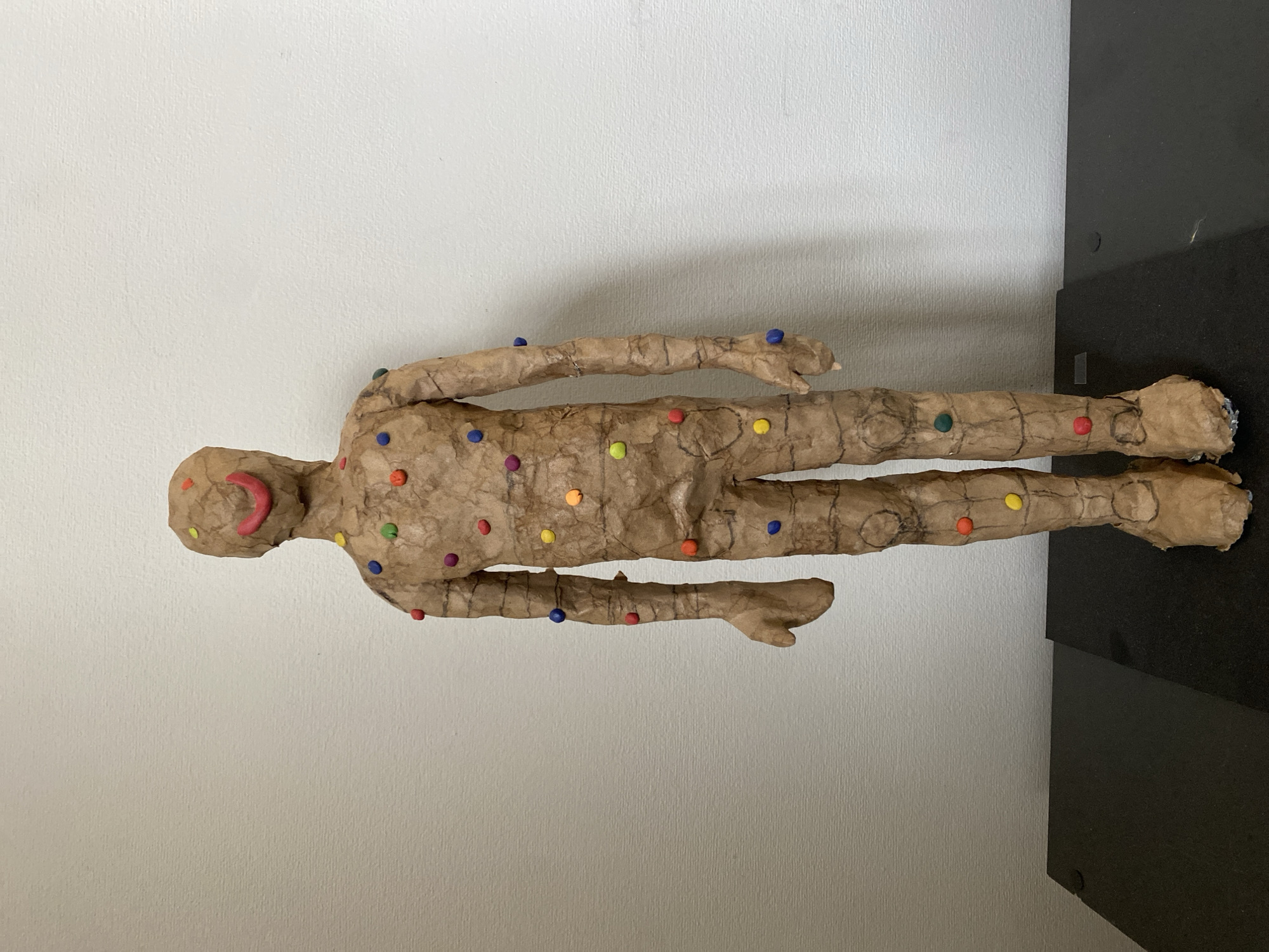 The image shows a sculpture of a human figure, lying horizontally on a surface. The sculpture appears to be made of a light-brown material, possibly paper or a similar substance. The figure is decorated with small, colorful dots. The background is a plain white wall and a black vertical surface, potentially a pedestal. There is no visual evidence in the image to indicate a specific location in Moscow, Russia.