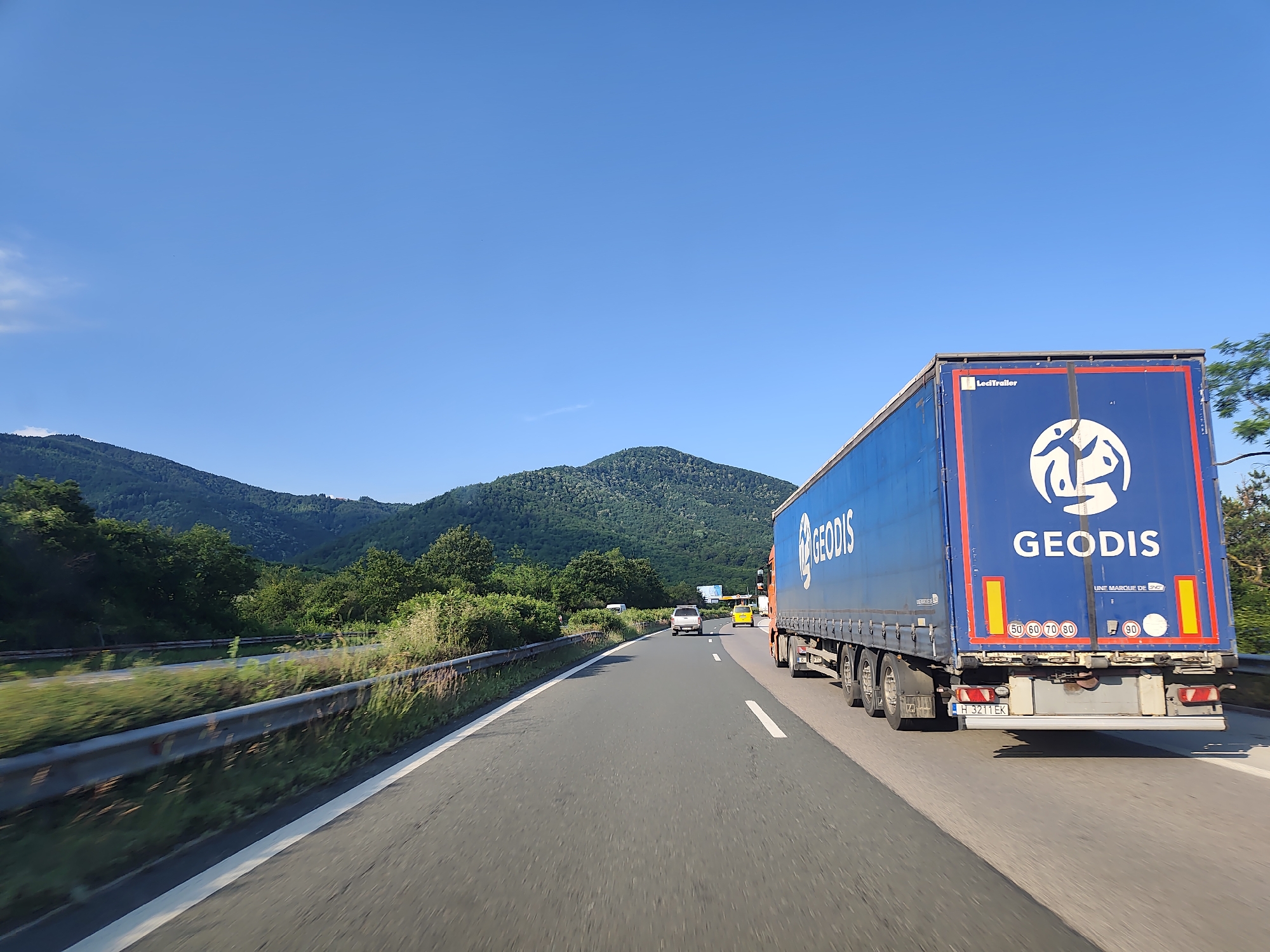 This is a daytime, outdoor, eye-level, medium shot of a blue semi-trailer truck with "GEODIS" written in white letters on its side and back. The truck is on a highway, driving alongside other vehicles, including a silver SUV and a yellow car. The highway is bordered by a metal guardrail and lush green trees and vegetation. In the background, rolling, tree-covered hills under a clear blue sky suggest a mountainous or hilly landscape. The sun is shining, indicating fair weather. The image appears to be taken from a moving vehicle, as suggested by the motion blur on the roadside vegetation. Visible text on the truck includes "GEODIS", "LeciTrailer", "UNE MARQUE DE G", "50 60 70 80", "90", and a license plate that reads "H 3211 EK". The overall scene depicts a common highway travel scenario.