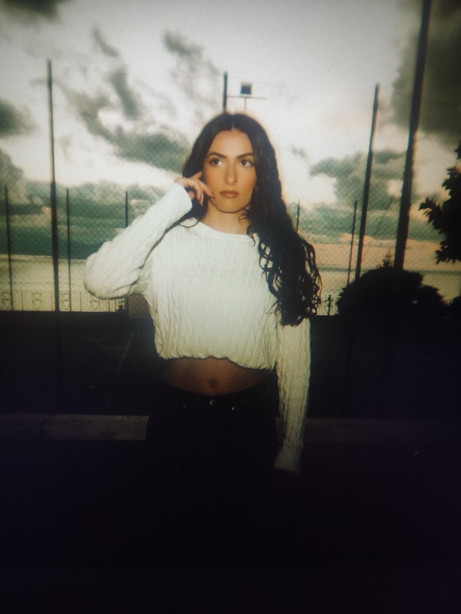 An outdoor portrait shows a young woman with long, dark, wavy hair and fair skin. She is wearing a white, cropped, cable-knit sweater and dark pants. The woman is standing in front of a fence and trees, with a cloudy sky in the background. She is looking off to the side, with her right hand touching her ear. The image appears to be taken at dusk.